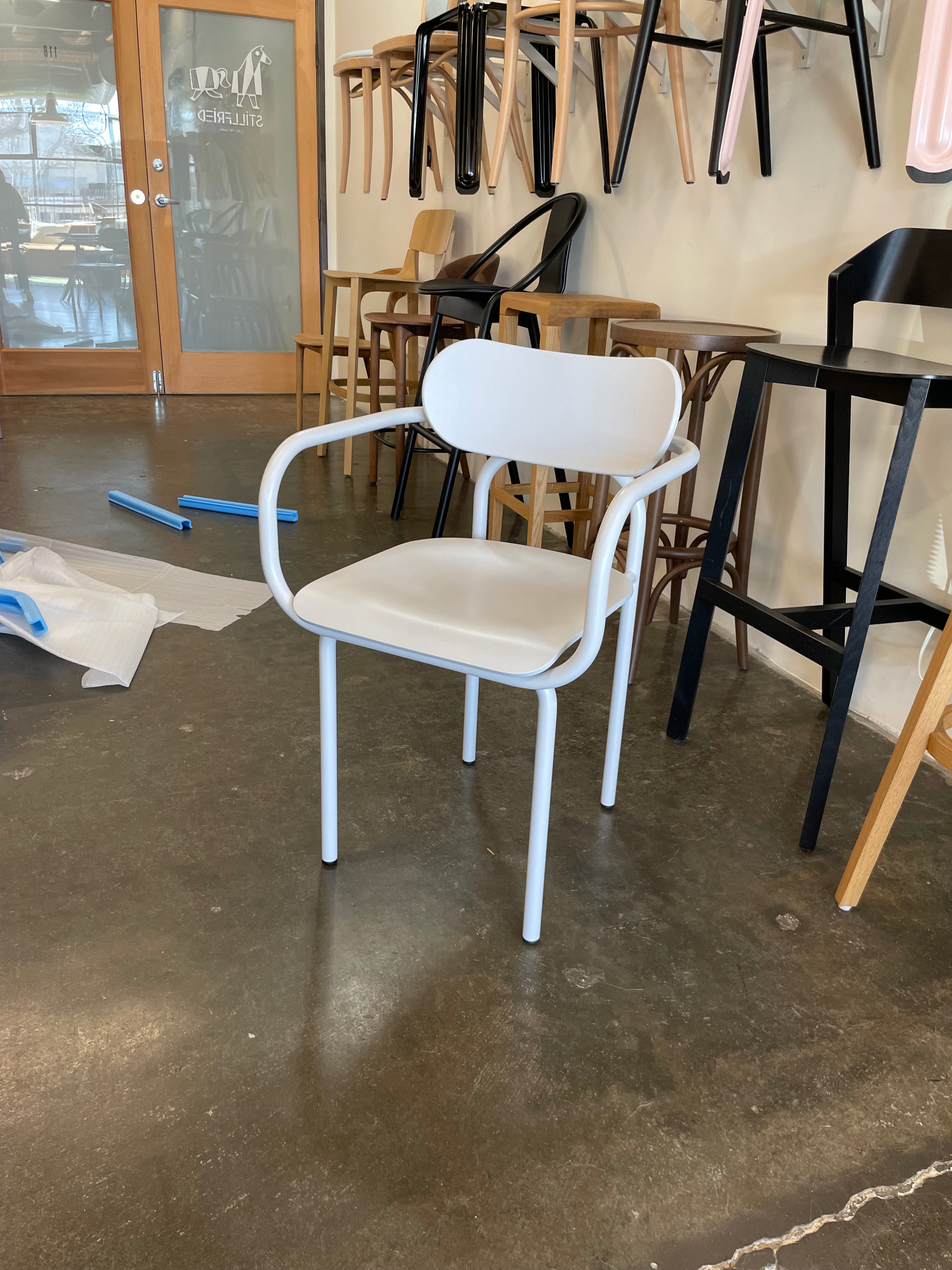 A modern white Victor Foxtrot Double U Armchair - Sale sits on a polished concrete floor in a showroom. Behind it, various chairs with iconic aesthetics hang on the wall, displaying multiple colors and styles. Packaging material is partially visible on the floor.