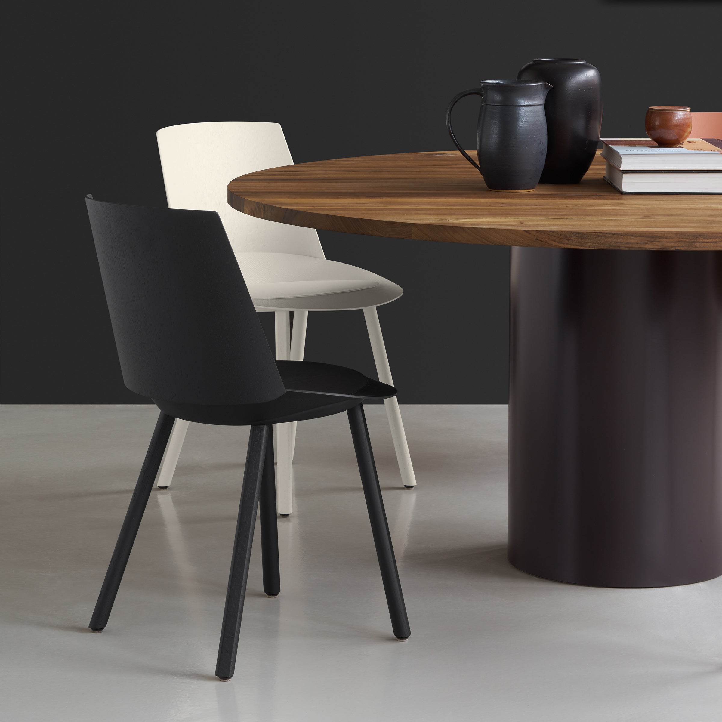 A modern dining scene features a round wooden table with a dark base, topped with books and ceramic vessels. The CH04 HOUDINI chairs by E15 add elegance around the table against the moody backdrop, showcasing a refined touch.
