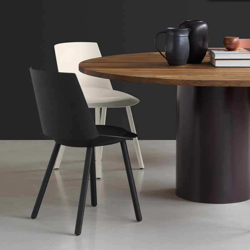 A modern dining scene features a round wooden table with a dark base, topped with books and ceramic vessels. The CH04 HOUDINI chairs by E15 add elegance around the table against the moody backdrop, showcasing a refined touch.
