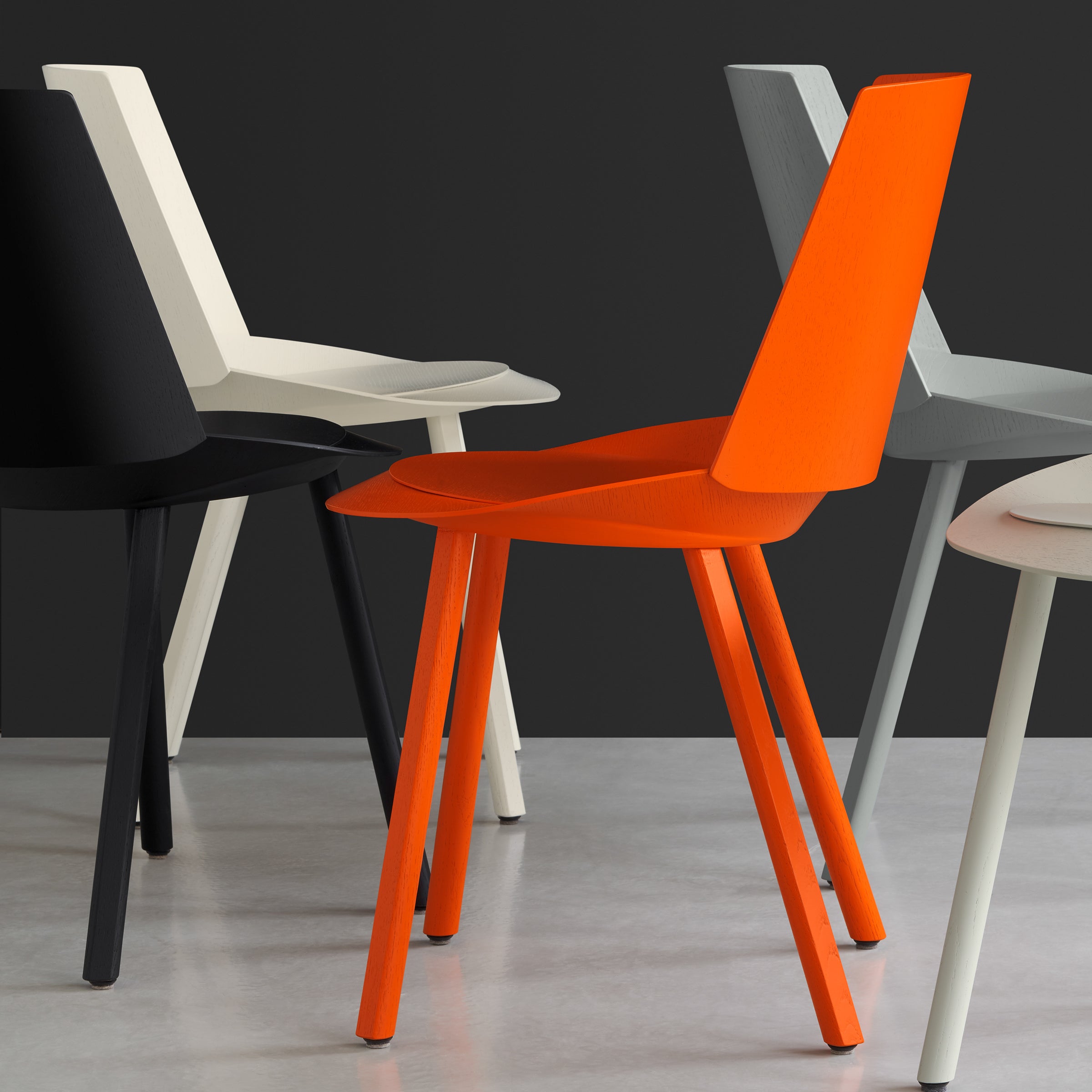Four modern chairs, featuring the striking E15 CH04 HOUDINI by Stefan Diez, showcase sleek, angular designs. They come in black, cream, gray, and a vibrant orange that stands out prominently against the dark backdrop on a light floor.