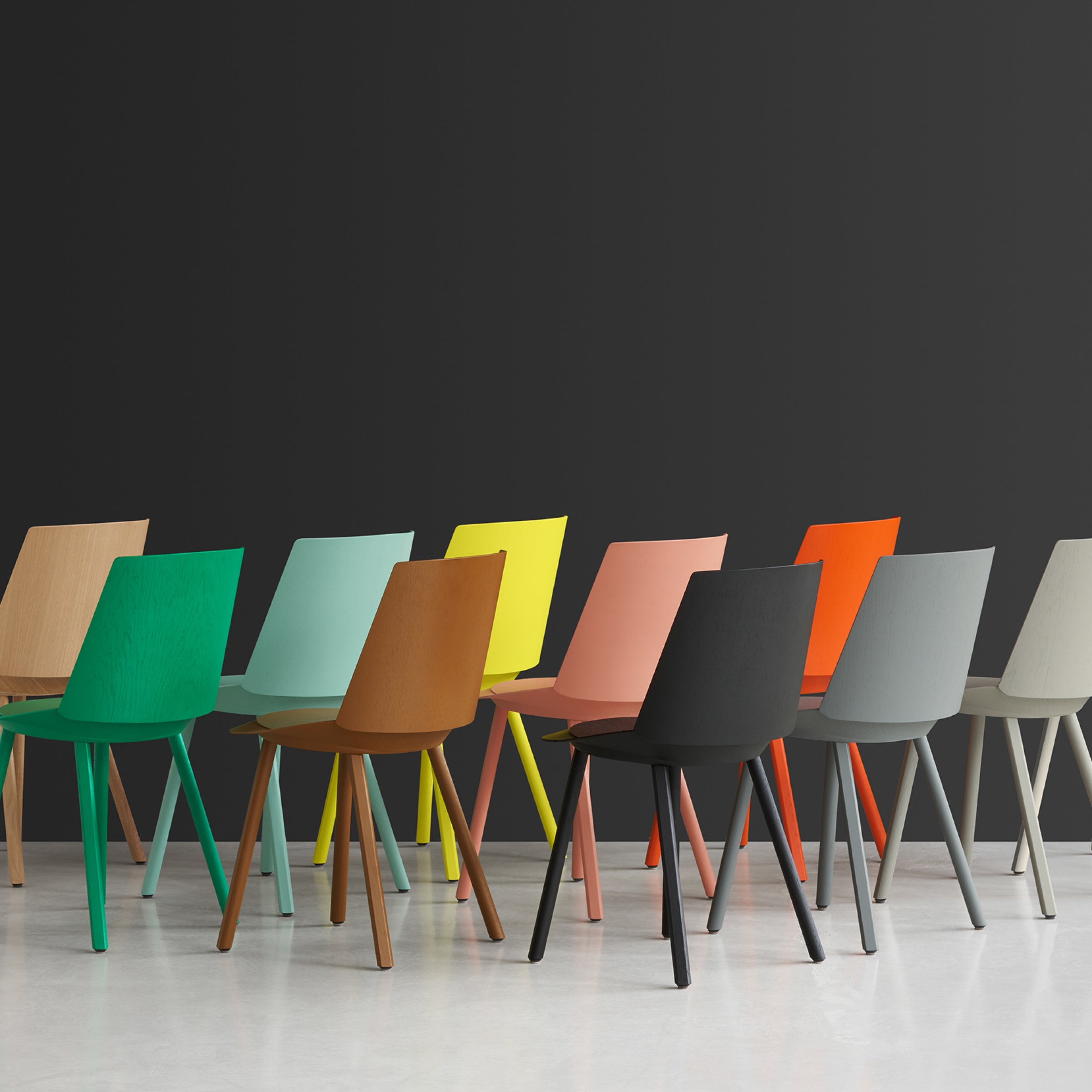 A row of colorful CH04 HOUDINI chairs by E15 in green, brown, yellow, pink, black, and gray vividly stand out against a dark gray wall with a light gray floor enhancing their vibrant contrast.