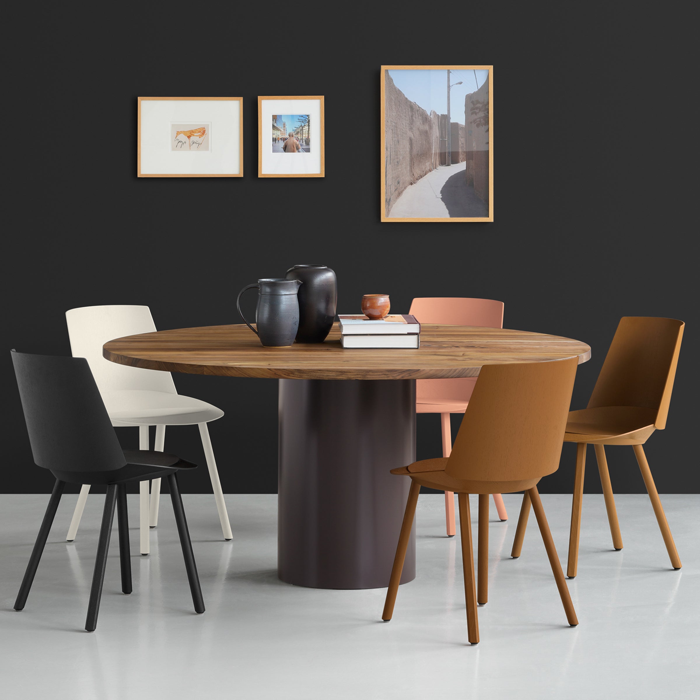 A round wooden table with a central dark base is encircled by five CH04 HOUDINI chairs in black, white, pink, and brown. Black teapots and mugs sit on the table. Three framed pictures adorn the dark wall behind it. White flooring highlights this E15 design by Stefan Diez.