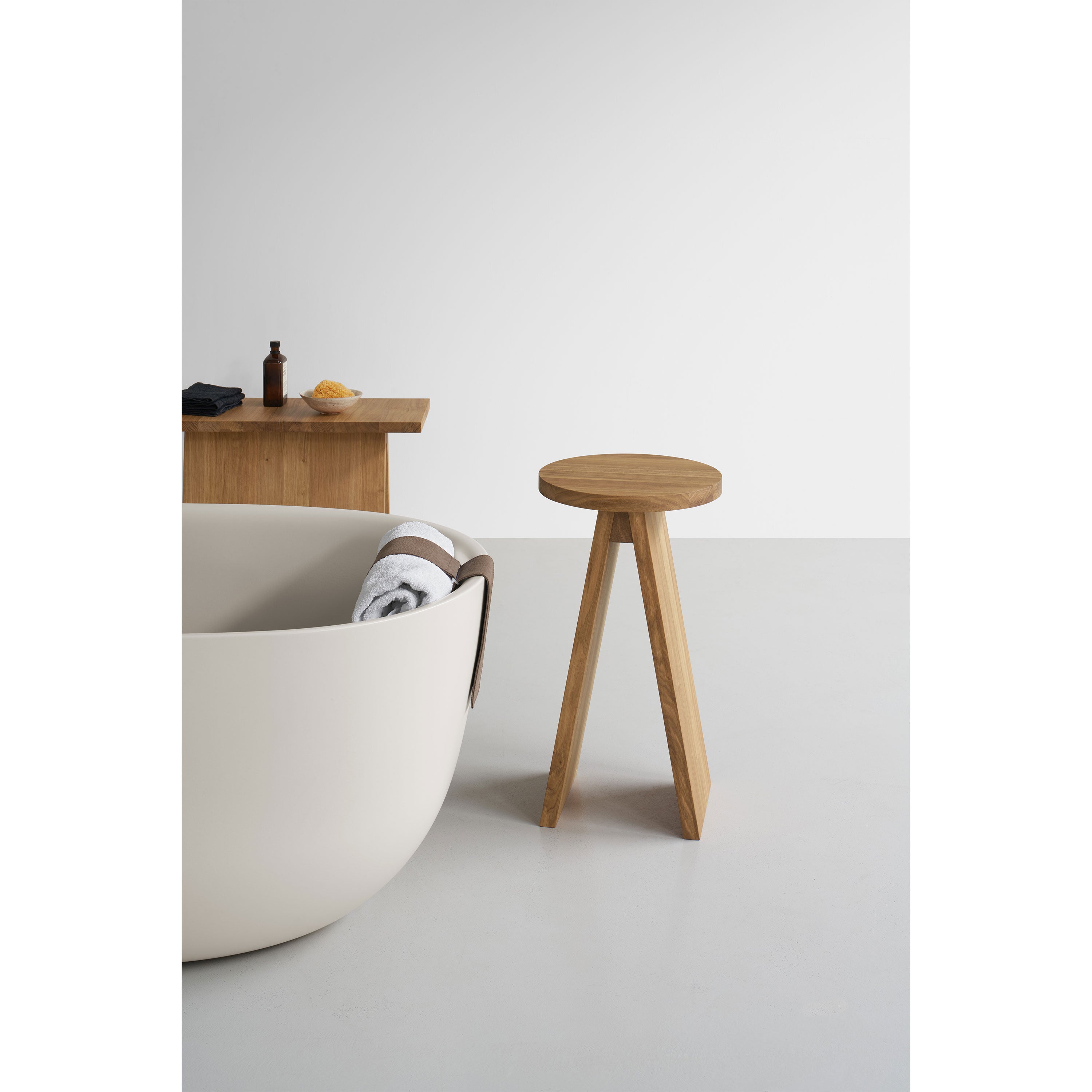 A minimalist bathroom showcases a white round bathtub with a towel and an E15 EK01/EK02 Stool in European oak beside it. A wooden table holds toiletries against the plain white wall, adding subtle elegance reminiscent of Stefan Diezs design ethos.