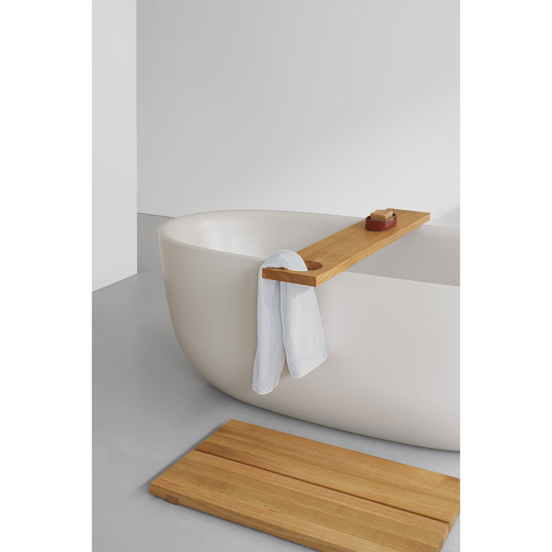 A modern bathroom showcases a white freestanding enamelled steel bathtub paired with the EK05 Bridge by E15—a light wood tray holding a soap dish and towel. A matching wooden bath mat lies on the gray floor, enhancing the minimalist vibe against crisp white walls.