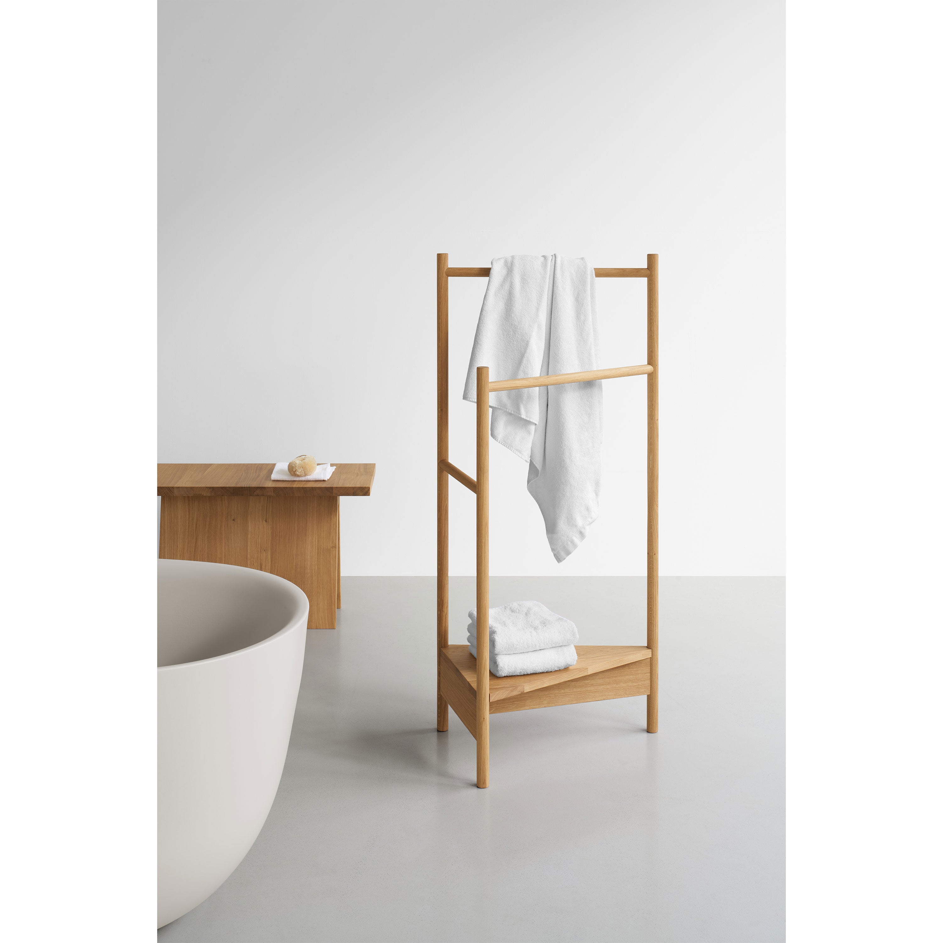 A minimalist bathroom showcases an EK04 Tripod towel rack by E15, crafted from solid European oak with white towels. A freestanding bathtub highlights the foreground, while a wooden bench with a small item completes the sleek, modern design in the background.