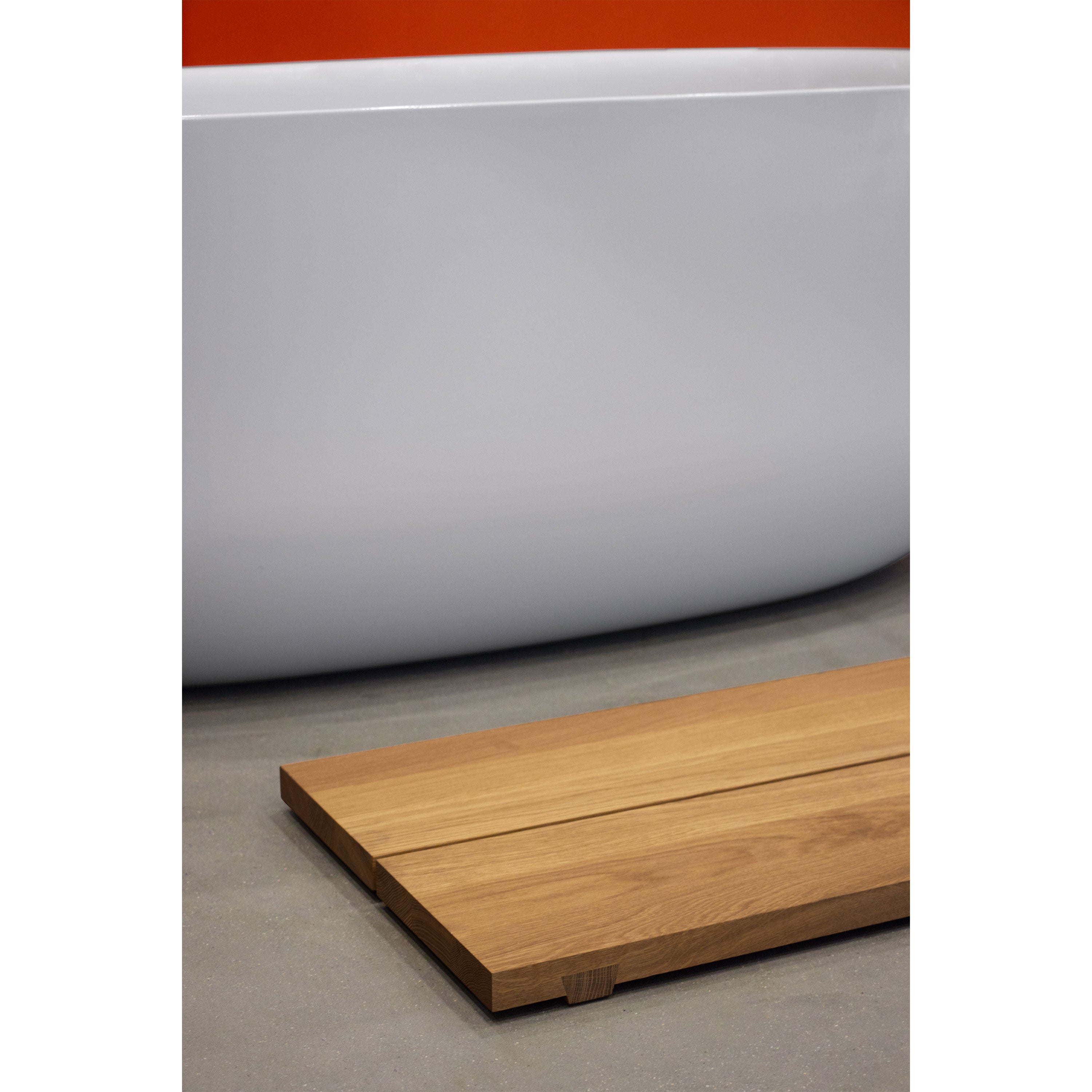 Close-up of a minimalist bathroom with a white bathtub against an orange wall. The EK06 Base bath mat by E15, crafted from European oak with traditional joinery, sits on the gray floor, enhancing the modern, clean aesthetic.