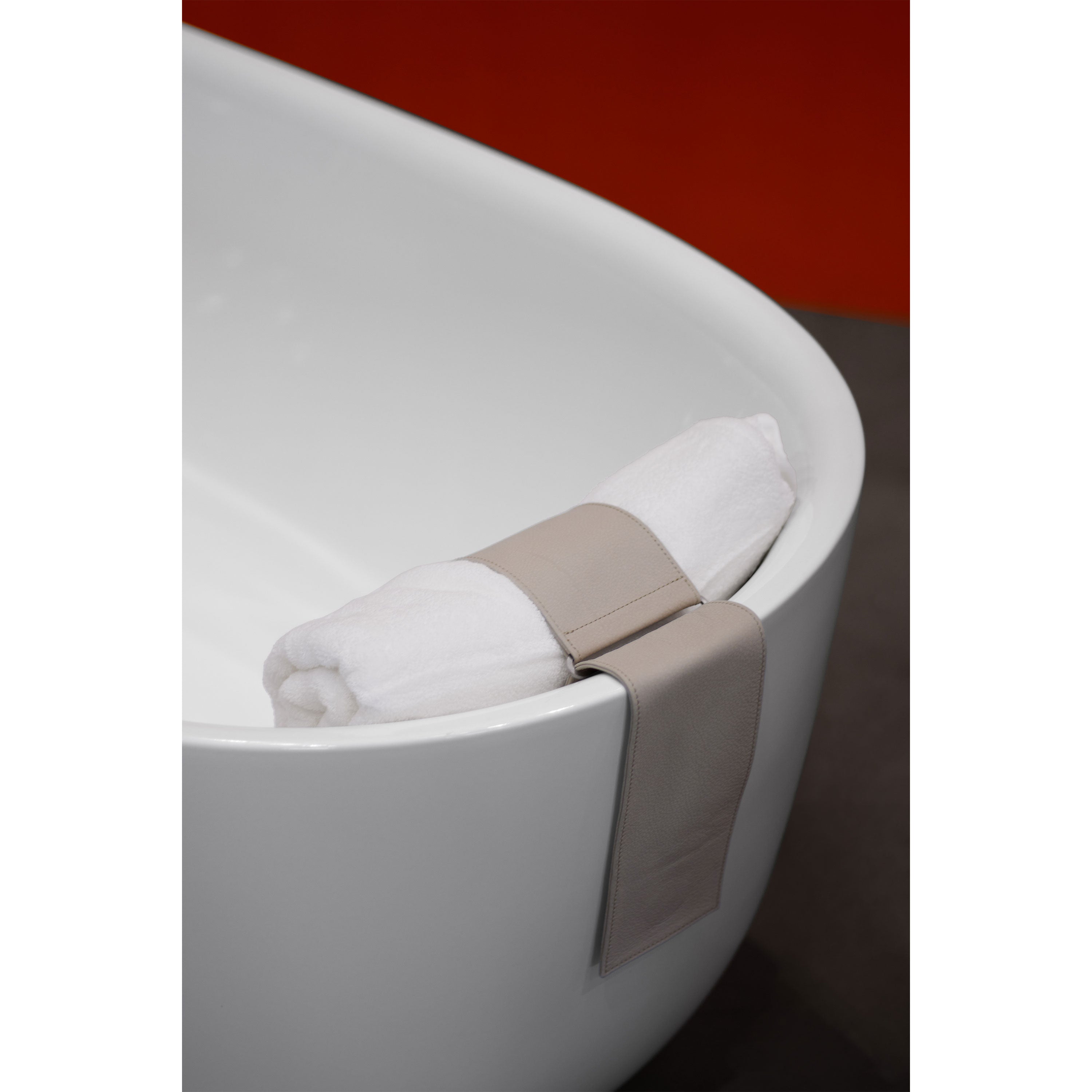 A white bathtub, likely a Kaldewei, displays a white towel rolled up and fastened with a beige leather strap on its rim. The setting includes a dark gray floor and part of a red wall, offering an elegant contrast. Featured product: EK08 Loop by E15.