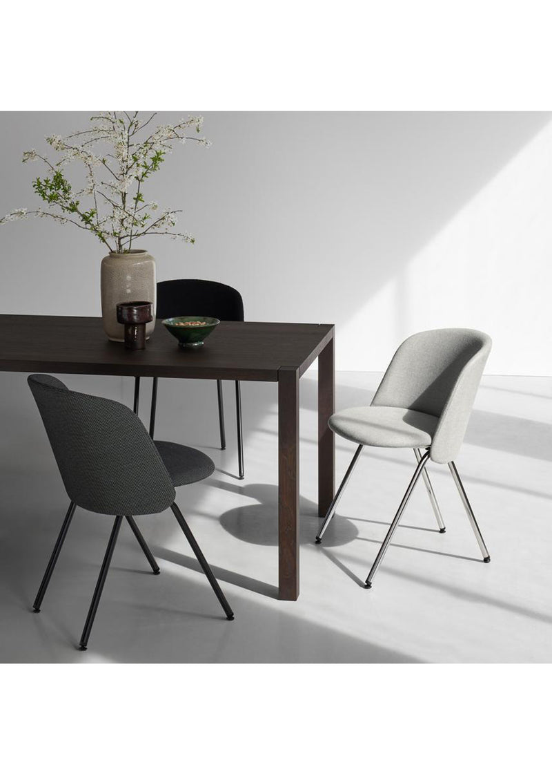 A modern dining area features a dark wood table paired with three CH07 EITHER Chairs by E15 in gray and black, exuding contemporary elegance. A vase with branches and a ceramic bowl enhance the tabletop, while sunlight creates gentle shadows on the gray floor and white wall.
