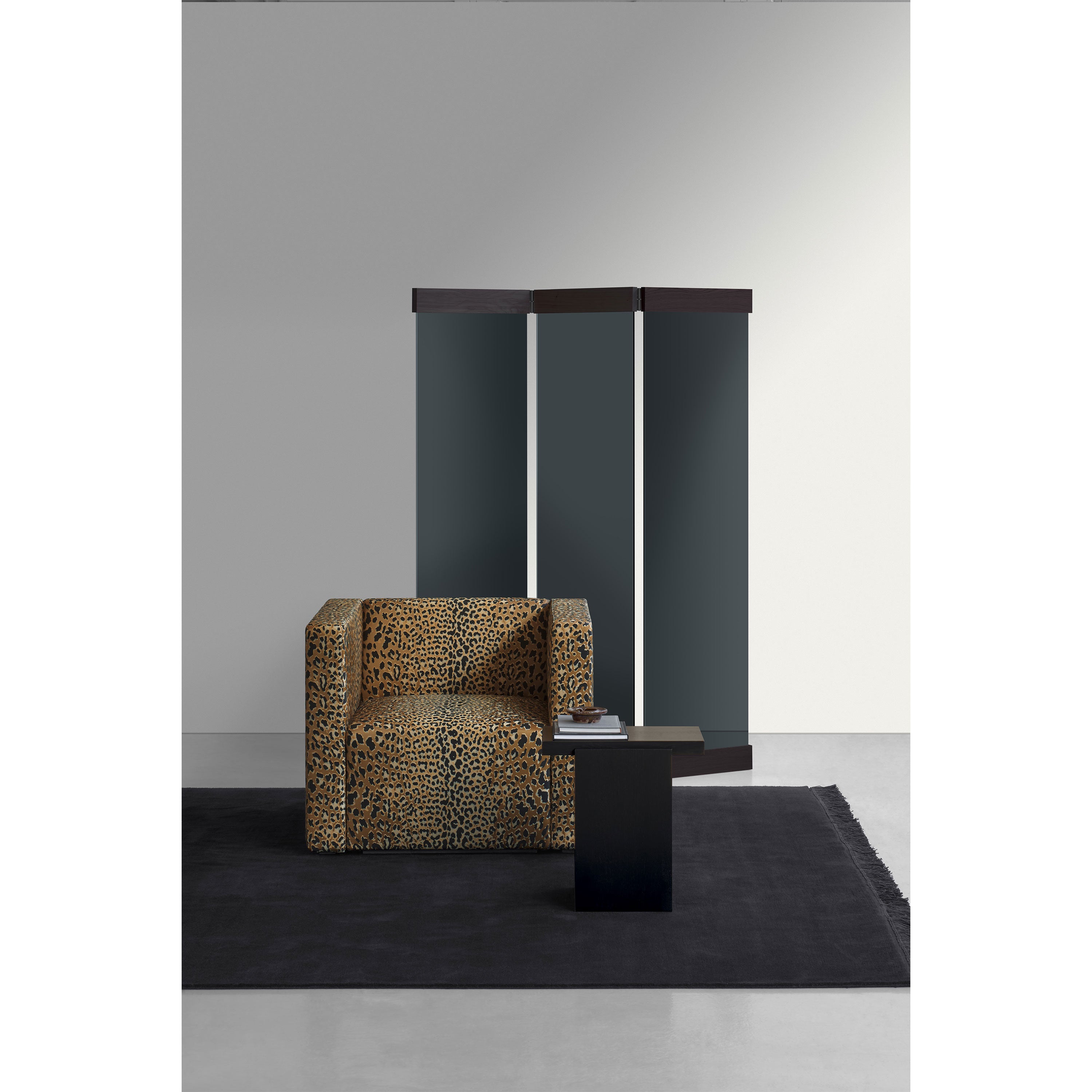 A modern interior includes a leopard print armchair beside a black, minimalist square side table on a dark rug. An Art Deco-inspired folding room divider featuring black and mirrored glass panels, the PV01 Scaena Paravent by E15, completes this minimalist aesthetic.