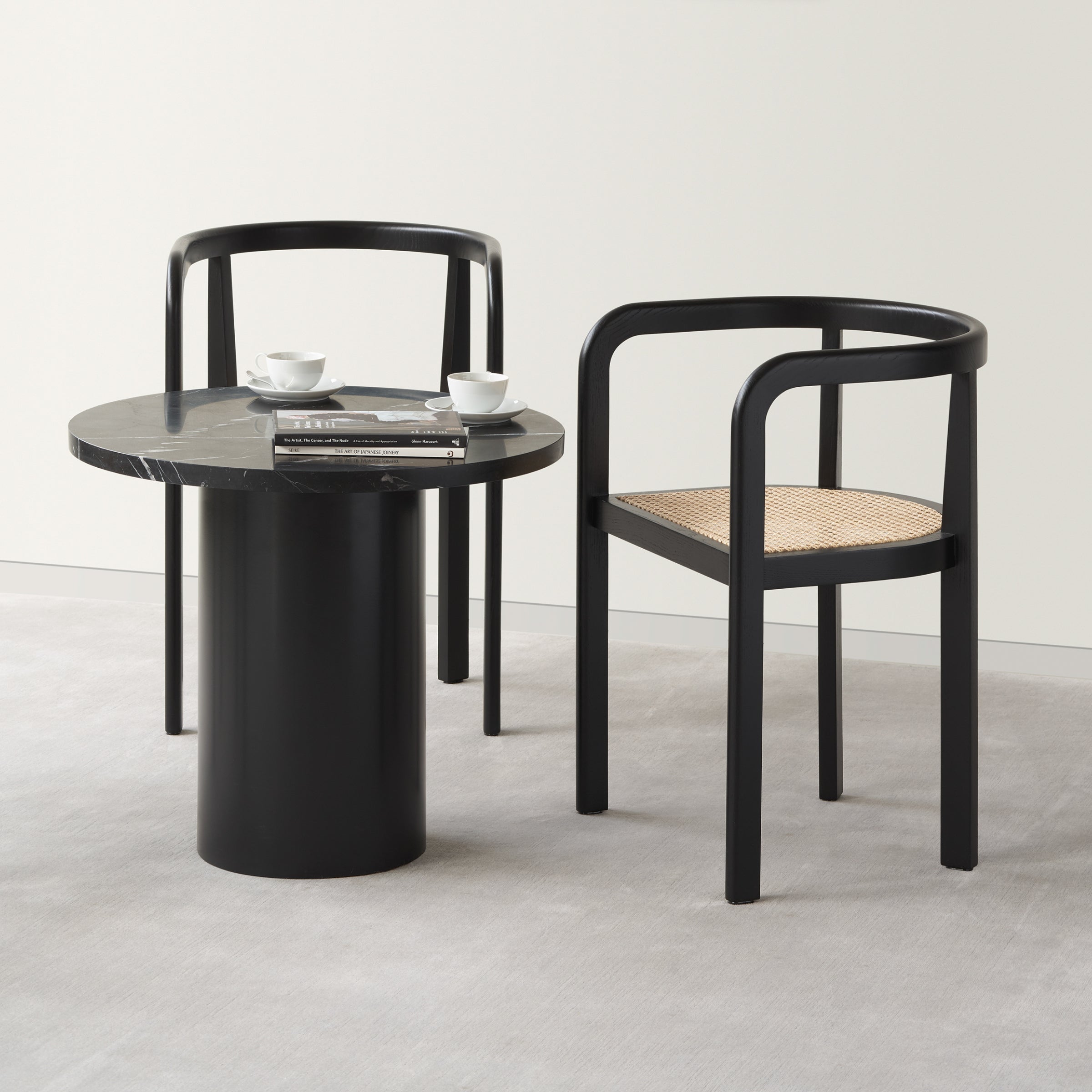 Two black RH01 Stuttgart chairs by E15, featuring curved arms and woven seats, surround a round black table. The table holds two cups and magazines, echoing Richard Herres design. The arrangement sits on a light gray carpet against a plain white wall.