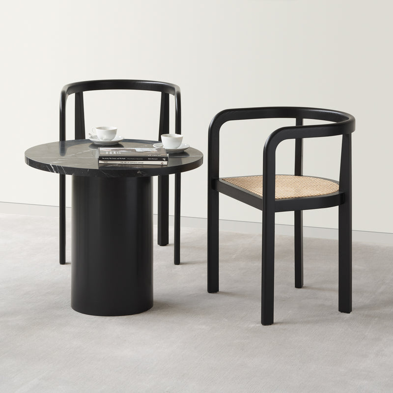 Two black RH01 Stuttgart chairs by E15, featuring curved arms and woven seats, surround a round black table. The table holds two cups and magazines, echoing Richard Herres design. The arrangement sits on a light gray carpet against a plain white wall.