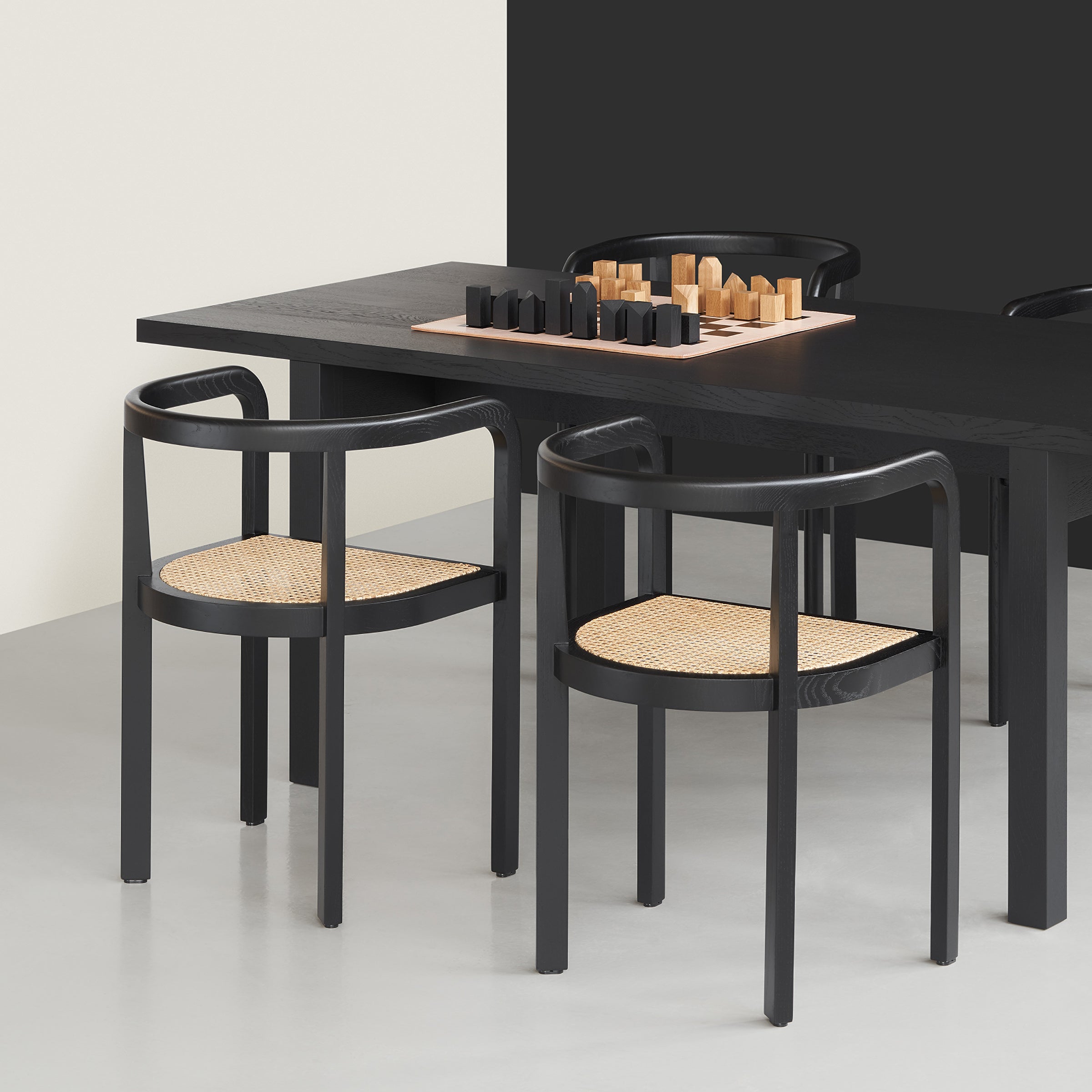 In a modern dining area, a black table and two RH01 Stuttgart chairs by E15 with woven rattan seats showcase exceptional wood craftsmanship. A chess set sits atop the table. Light and dark walls in the background enhance the Richard Herre-inspired contemporary design.