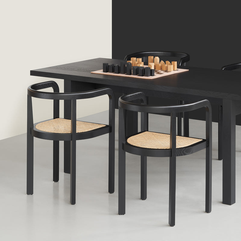 In a modern dining area, a black table and two RH01 Stuttgart chairs by E15 with woven rattan seats showcase exceptional wood craftsmanship. A chess set sits atop the table. Light and dark walls in the background enhance the Richard Herre-inspired contemporary design.