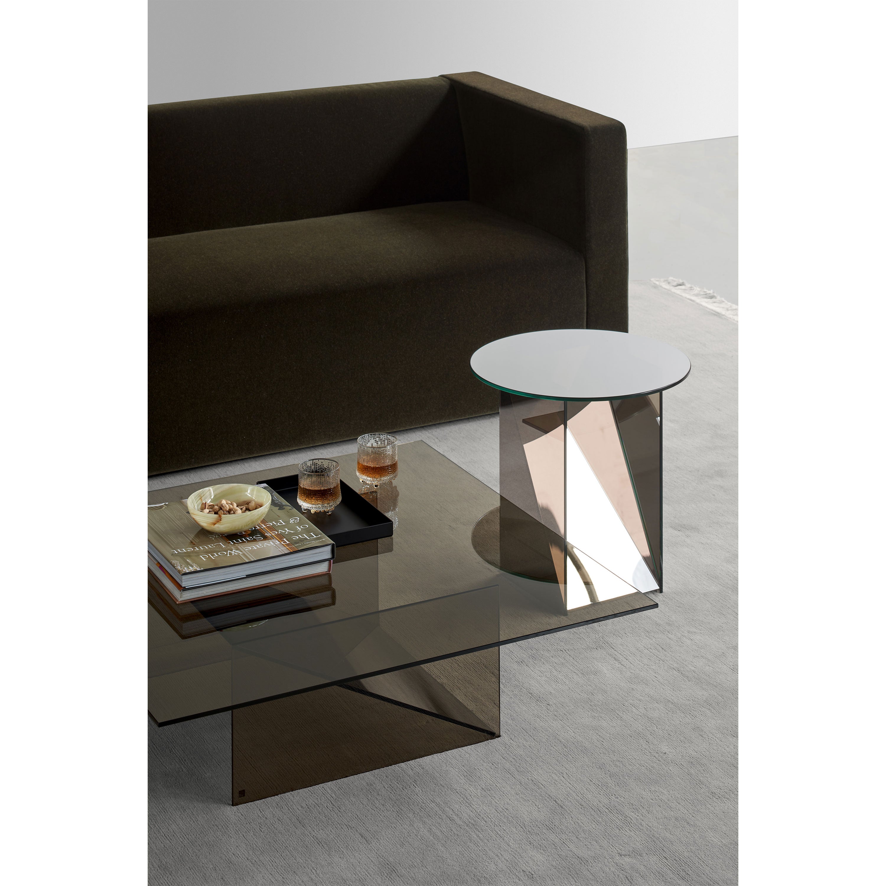 A modern living room features a dark sofa, an E15 CT11 Kaisa sleek glass coffee table by Annabelle Klute, and a small mirrored side table. The coffee table holds a book, a decorative tray with three glass items, and snacks in a bowl, all set on a light-colored floor.