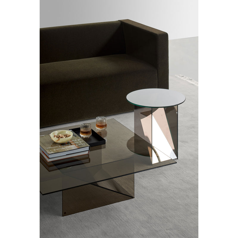 A modern living room features a dark sofa, an E15 CT11 Kaisa sleek glass coffee table by Annabelle Klute, and a small mirrored side table. The coffee table holds a book, a decorative tray with three glass items, and snacks in a bowl, all set on a light-colored floor.