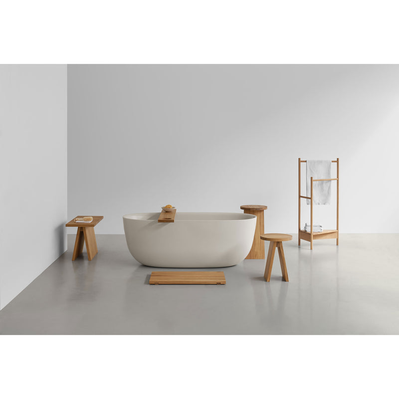 A minimalist bathroom showcases a white freestanding bathtub and a solid European oak towel rack. Wooden accents include the E15 EK04 Tripod stool, side table, and slatted mat. A soap bar sits on the side table, complementing the elegant smooth gray floor.