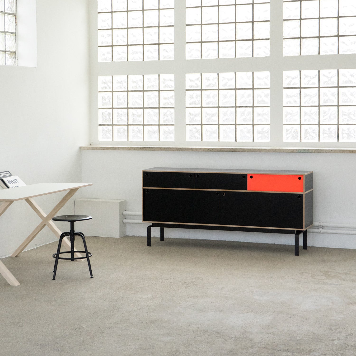 A minimalist room with white walls and glass blocks has a sleek white table and black stool. The modern Moormann Egal Sideboard, featuring a single orange drawer, adds contrast as its sliding doors blend with the industrial look of the concrete floor.