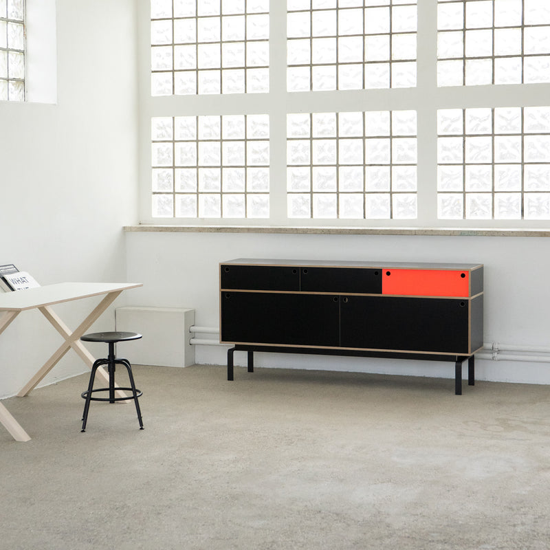 A minimalist room with white walls and glass blocks has a sleek white table and black stool. The modern Moormann Egal Sideboard, featuring a single orange drawer, adds contrast as its sliding doors blend with the industrial look of the concrete floor.