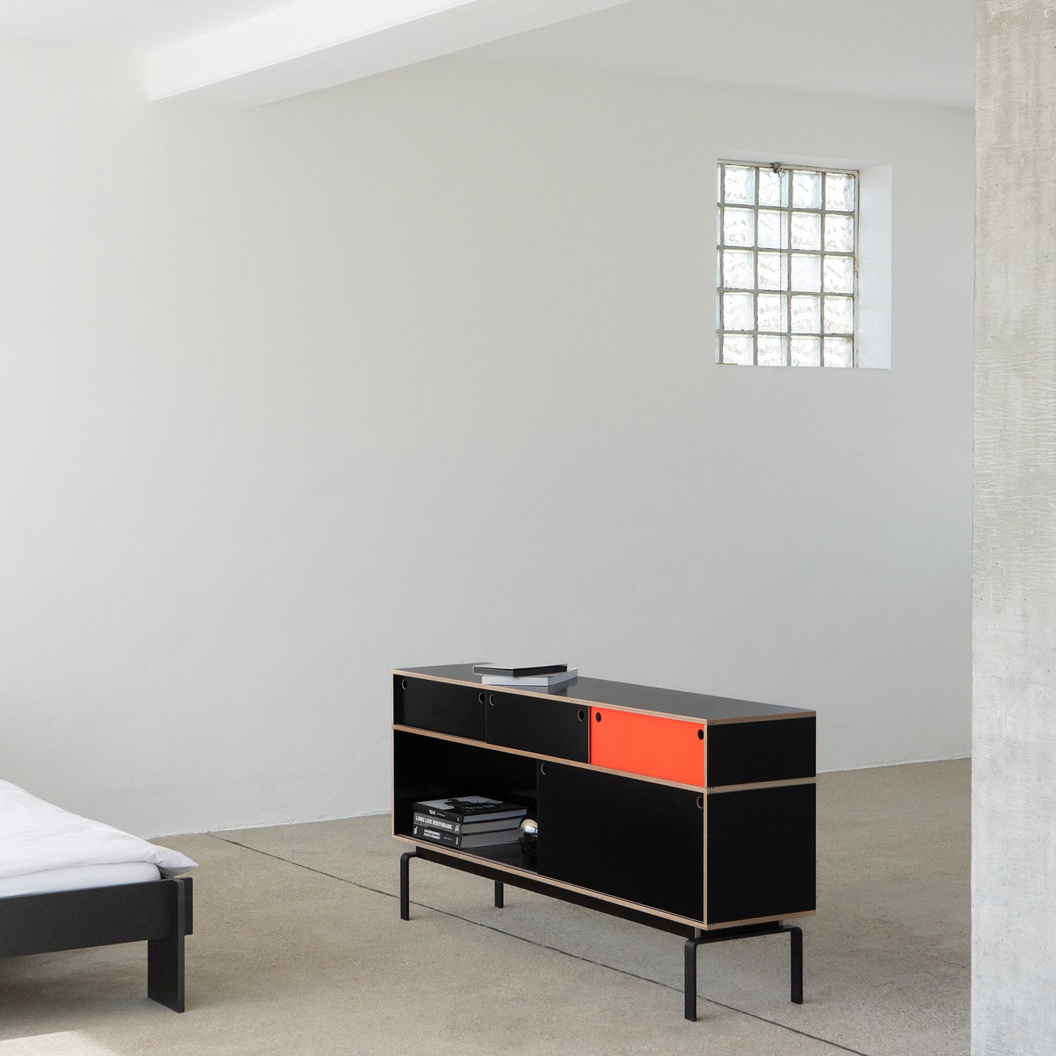In a minimalist room, a Moormann Egal sideboard with sleek sliding doors and an orange panel stands against a white wall. A bed with white linens peeks in from the left, and a square glass block window softly lights the light gray floor.