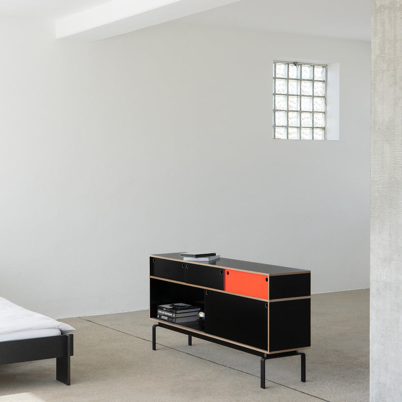 In a minimalist room, a Moormann Egal sideboard with sleek sliding doors and an orange panel stands against a white wall. A bed with white linens peeks in from the left, and a square glass block window softly lights the light gray floor.