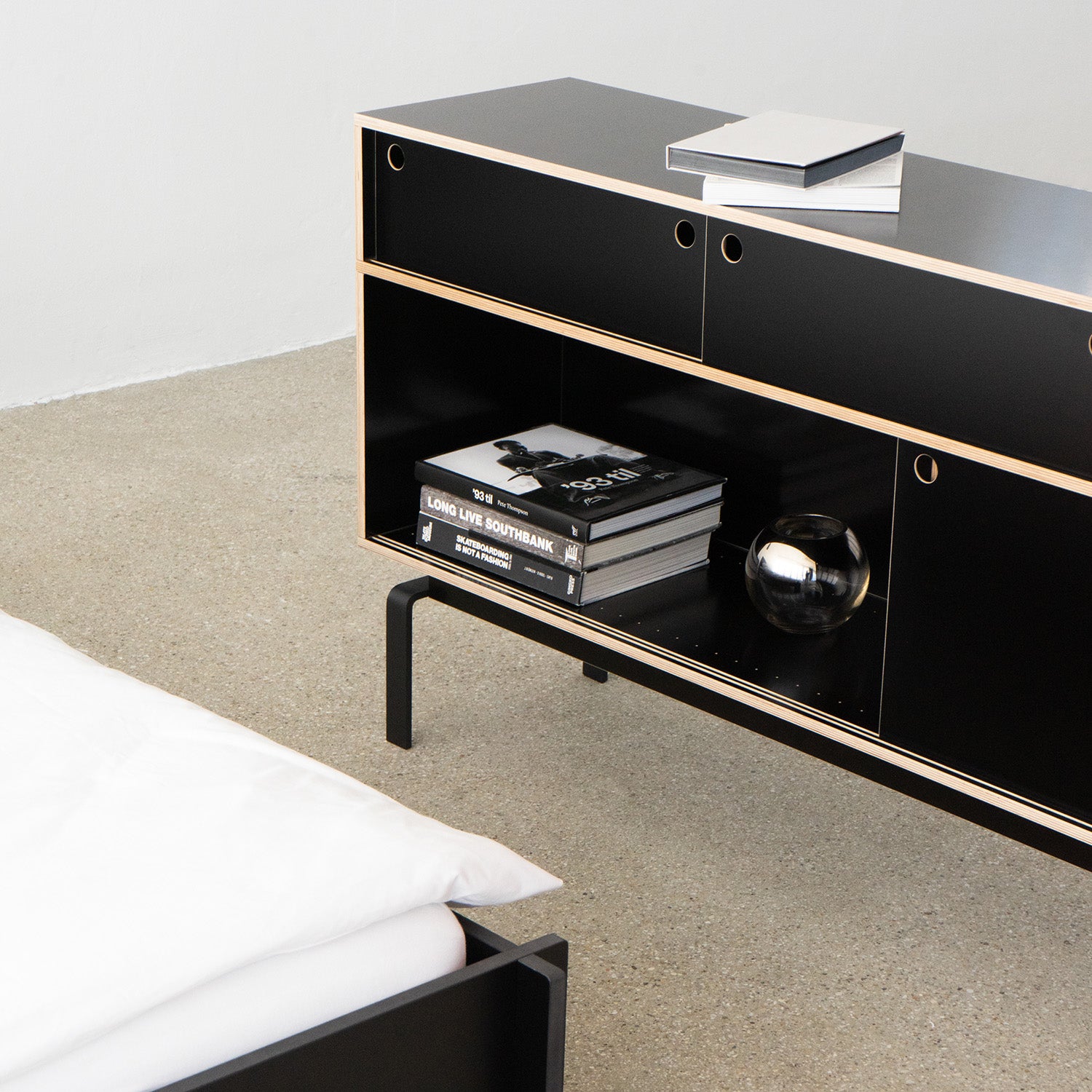 A minimalist room showcases the Moormann Egal Sideboard, featuring open shelves with stacked books and a shiny metallic sphere. A white bed is partially visible in front of a neutral-tone carpet and plain white wall.