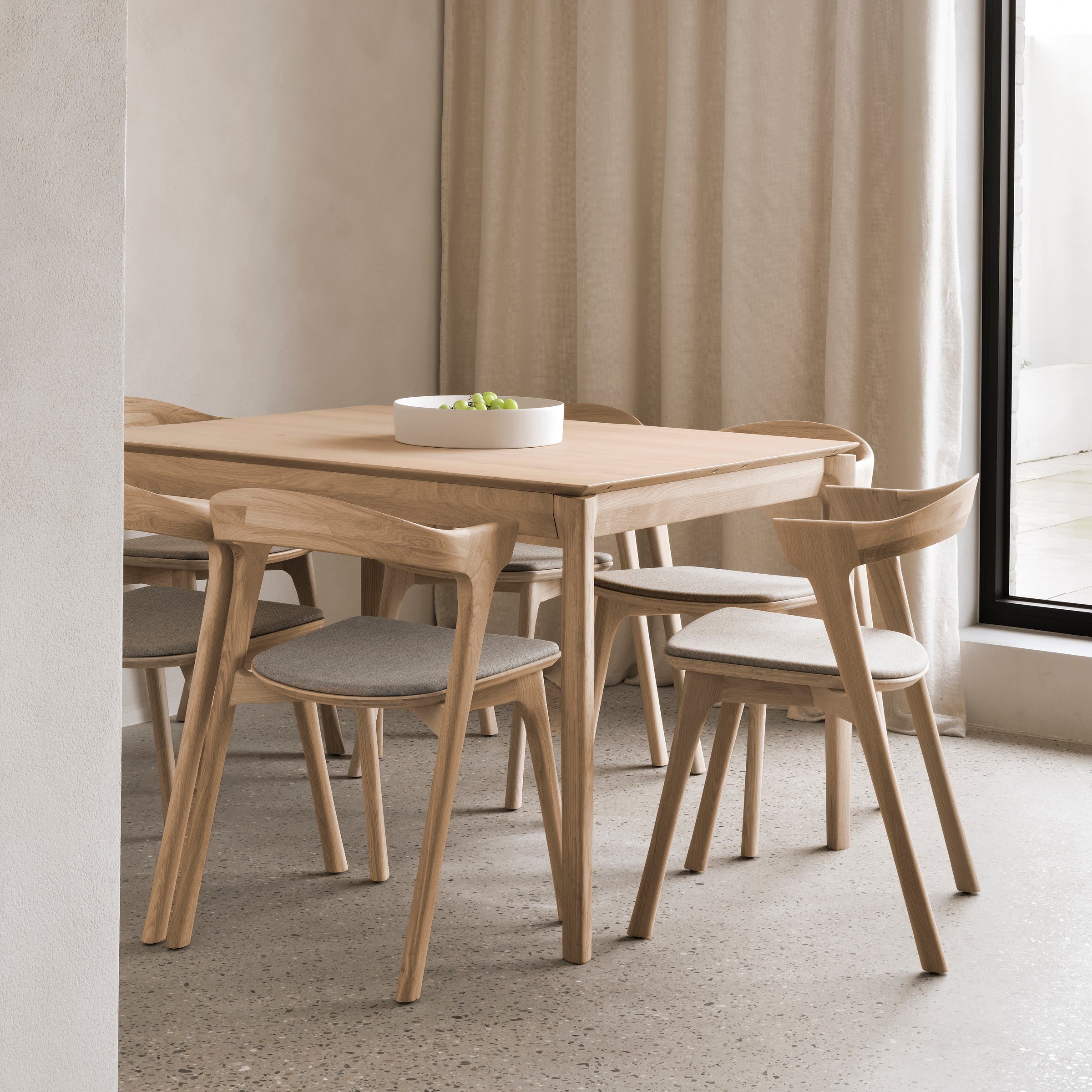 A minimalist dining area showcases the Ethnicraft Bok Extendable Dining Table (Rectangular), four cushioned wooden chairs, a white bowl of green apples, neutral walls, soft curtains, and a large window welcoming natural light.