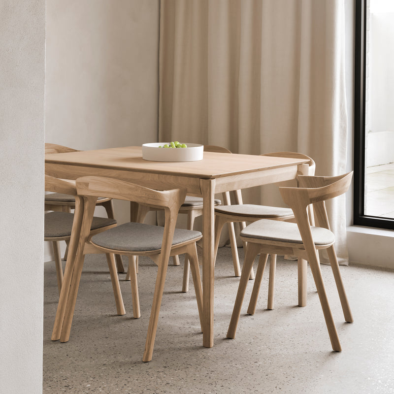 A minimalist dining area showcases the Ethnicraft Bok Extendable Dining Table (Rectangular), four cushioned wooden chairs, a white bowl of green apples, neutral walls, soft curtains, and a large window welcoming natural light.