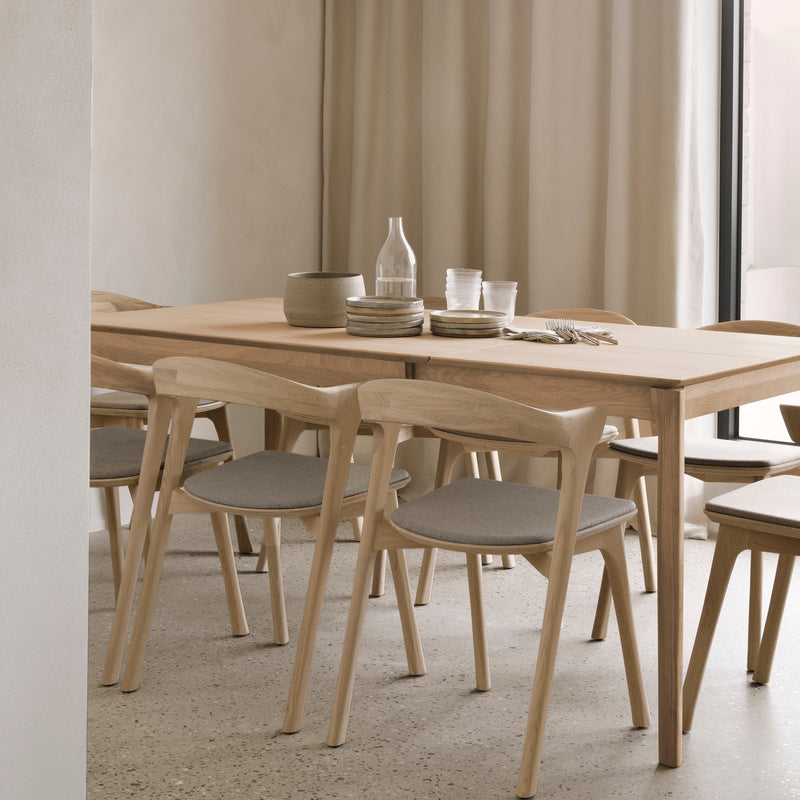 A minimalist dining room showcases the Ethnicraft Bok Extendable Dining Table – Rectangular with matching chairs and gray cushions. Stacked plates, glasses, a carafe, and a ceramic bowl sit atop the table; neutral curtains and walls add calm.