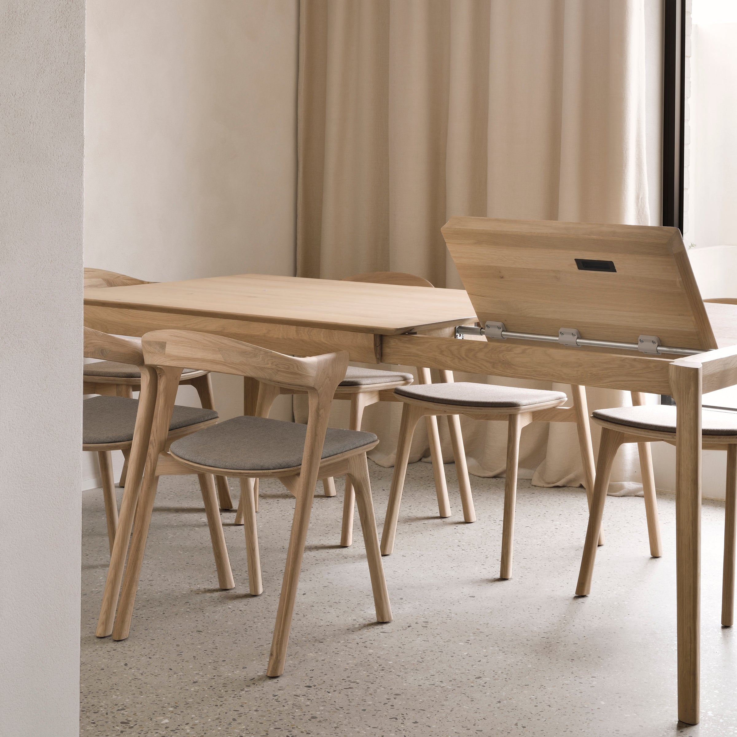 A minimalist dining area features light wooden chairs and the Ethnicraft Bok Extendable Dining Table - Rectangular, partially opened. Beige curtains and a polished concrete floor complete the contemporary look.