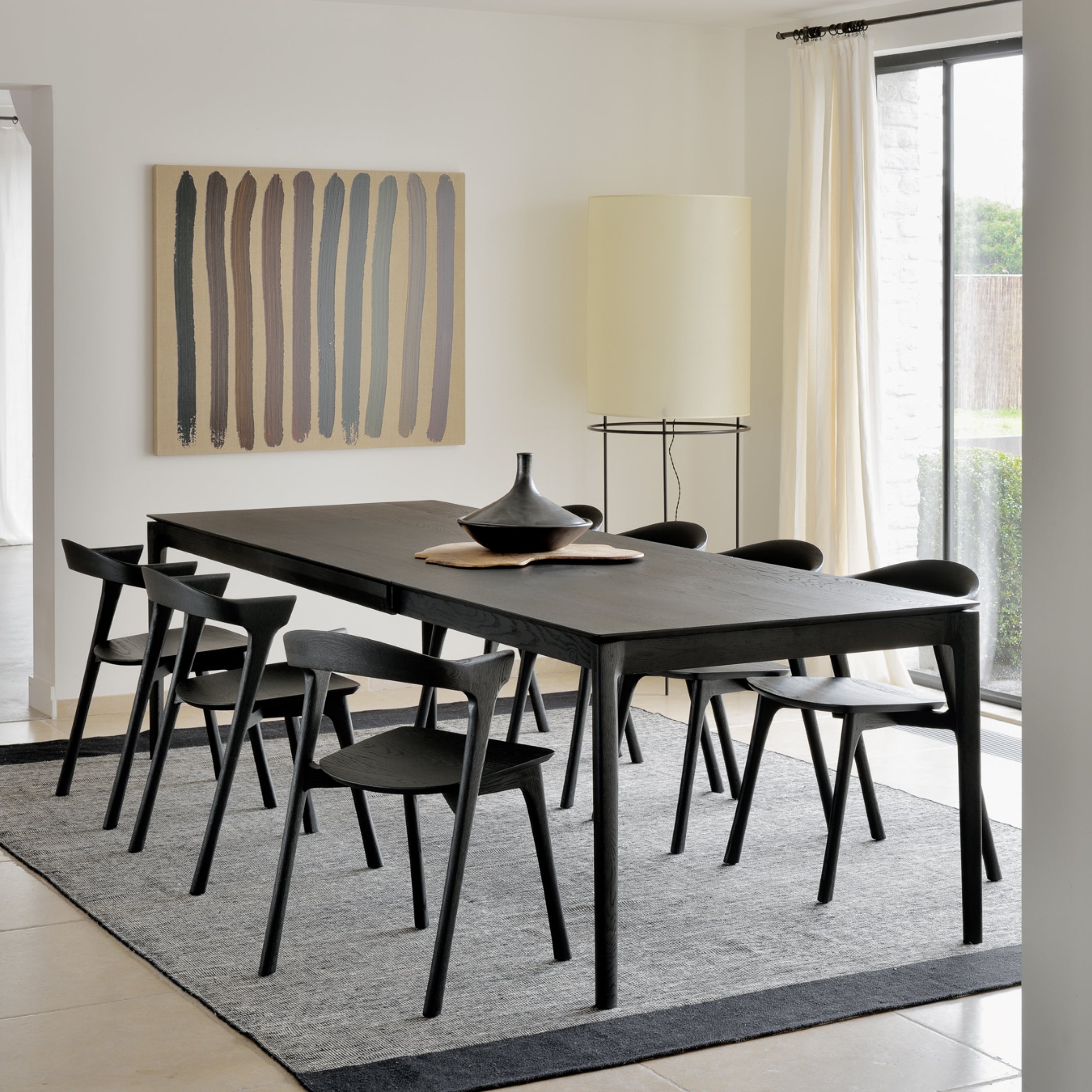 A modern dining room with the Ethnicraft Bok Extendable Dining Table, six black chairs, a gray rug, minimalist vase centerpiece, striped abstract wall art, a tall yellow lamp, and large windows with flowing white curtains.