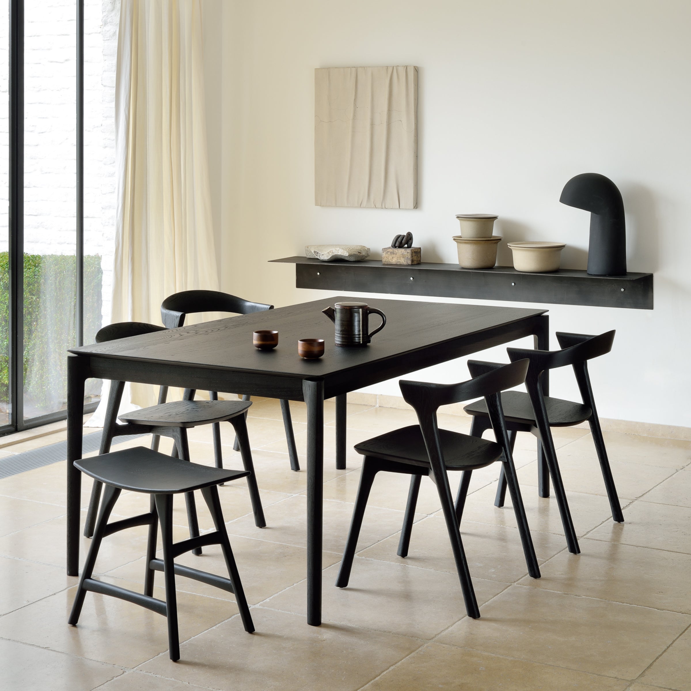 A modern dining area showcases Ethnicraft's Bok Extendable Dining Table in black, surrounded by five matching chairs, two sleek stools, ceramic bowls, and a sculptural accent on a floating shelf against a white wall.