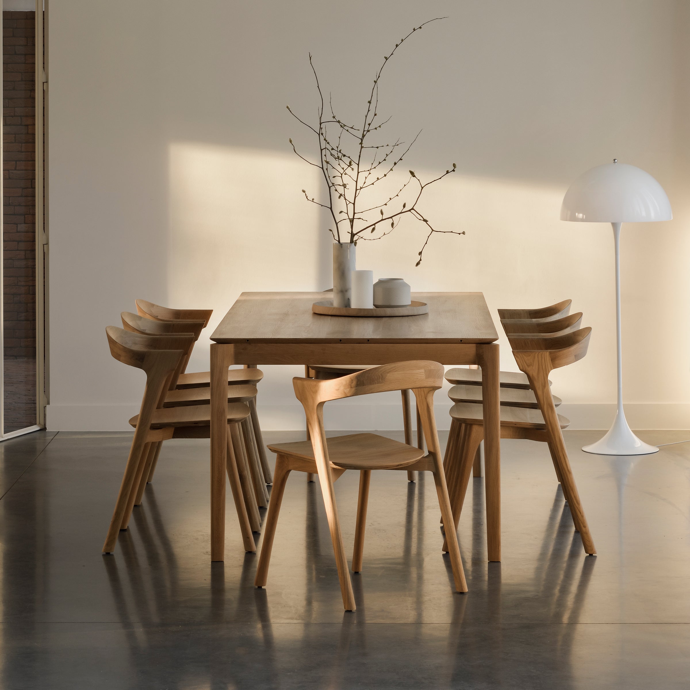A minimalist dining room features the Ethnicraft Bok Extendable Dining Table (Rectangular) with six wooden chairs, a white round-based floor lamp, and bare branches in a vase. Warm sunlight streams in, casting soft shadows on neutral walls and a polished floor.