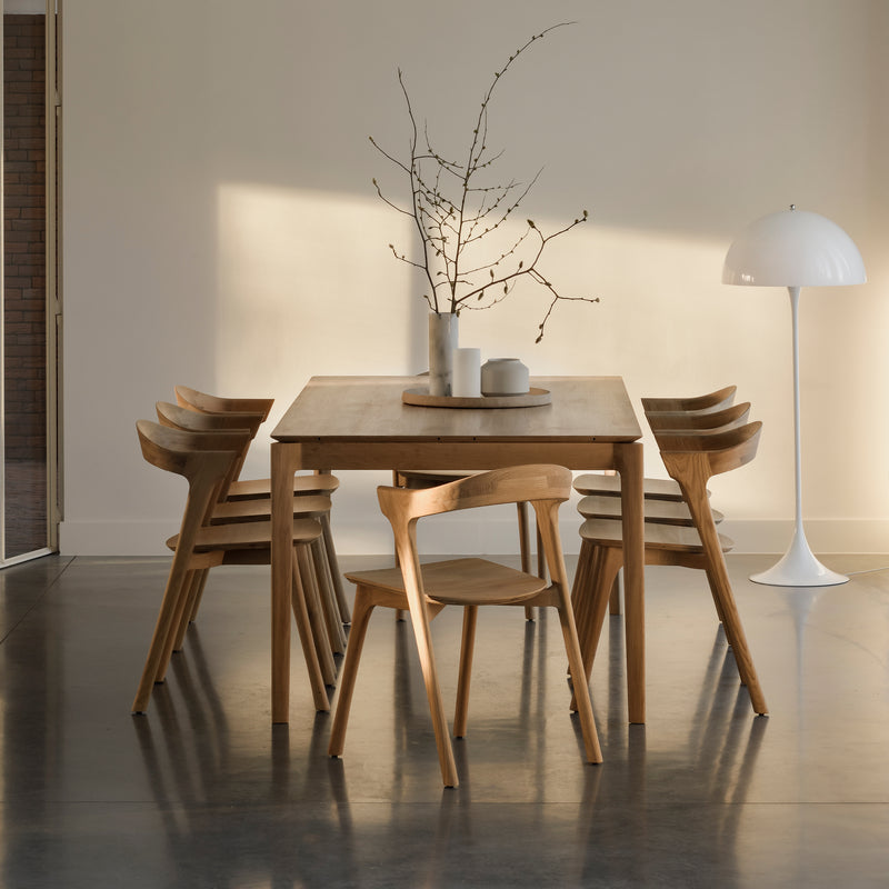A minimalist dining room features the Ethnicraft Bok Extendable Dining Table (Rectangular) with six wooden chairs, a white round-based floor lamp, and bare branches in a vase. Warm sunlight streams in, casting soft shadows on neutral walls and a polished floor.