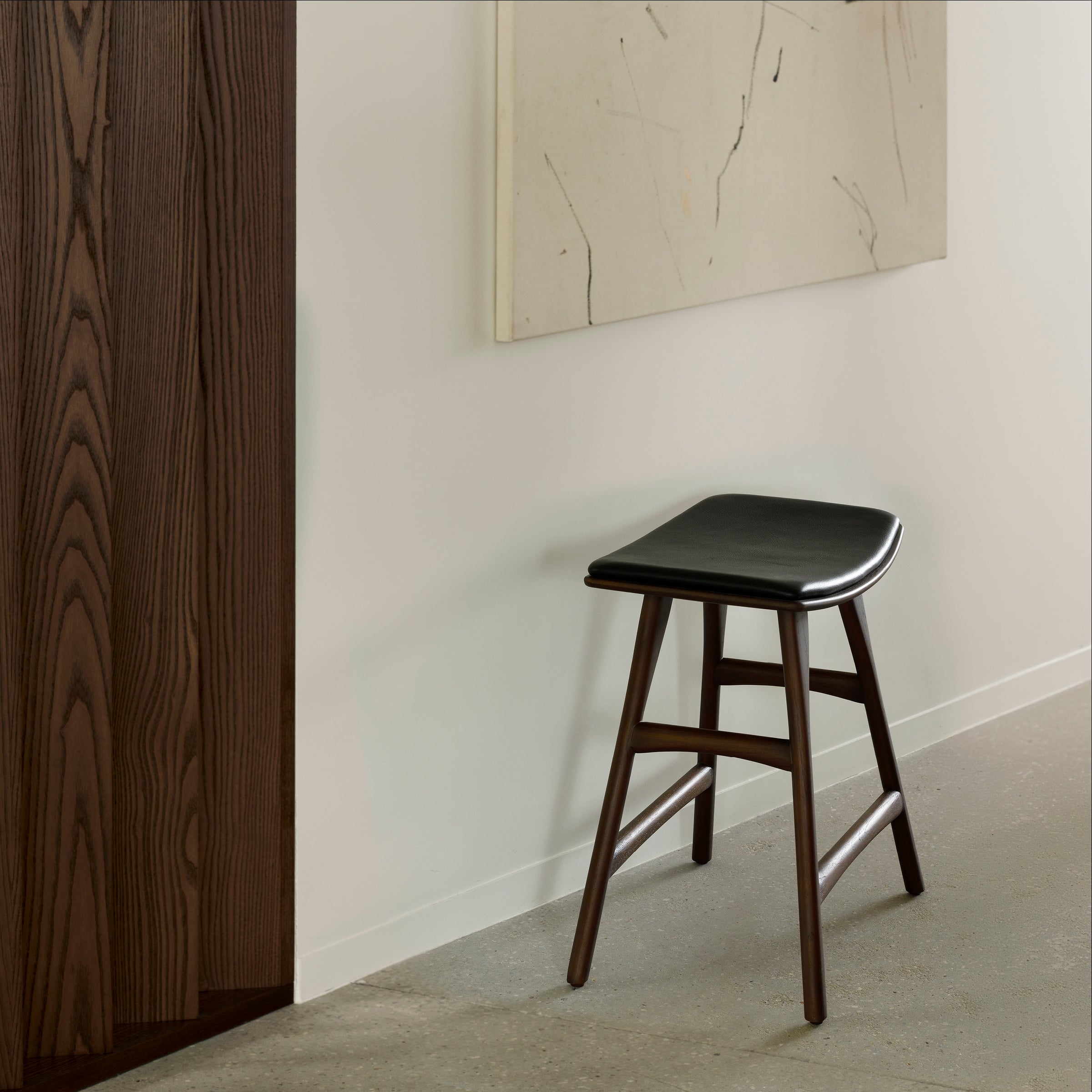 An Ethnicraft Osso Stool in oak with a black seat rests against a white wall below an abstract painting, set in a minimalist room with concrete floors and a dark wood panel.