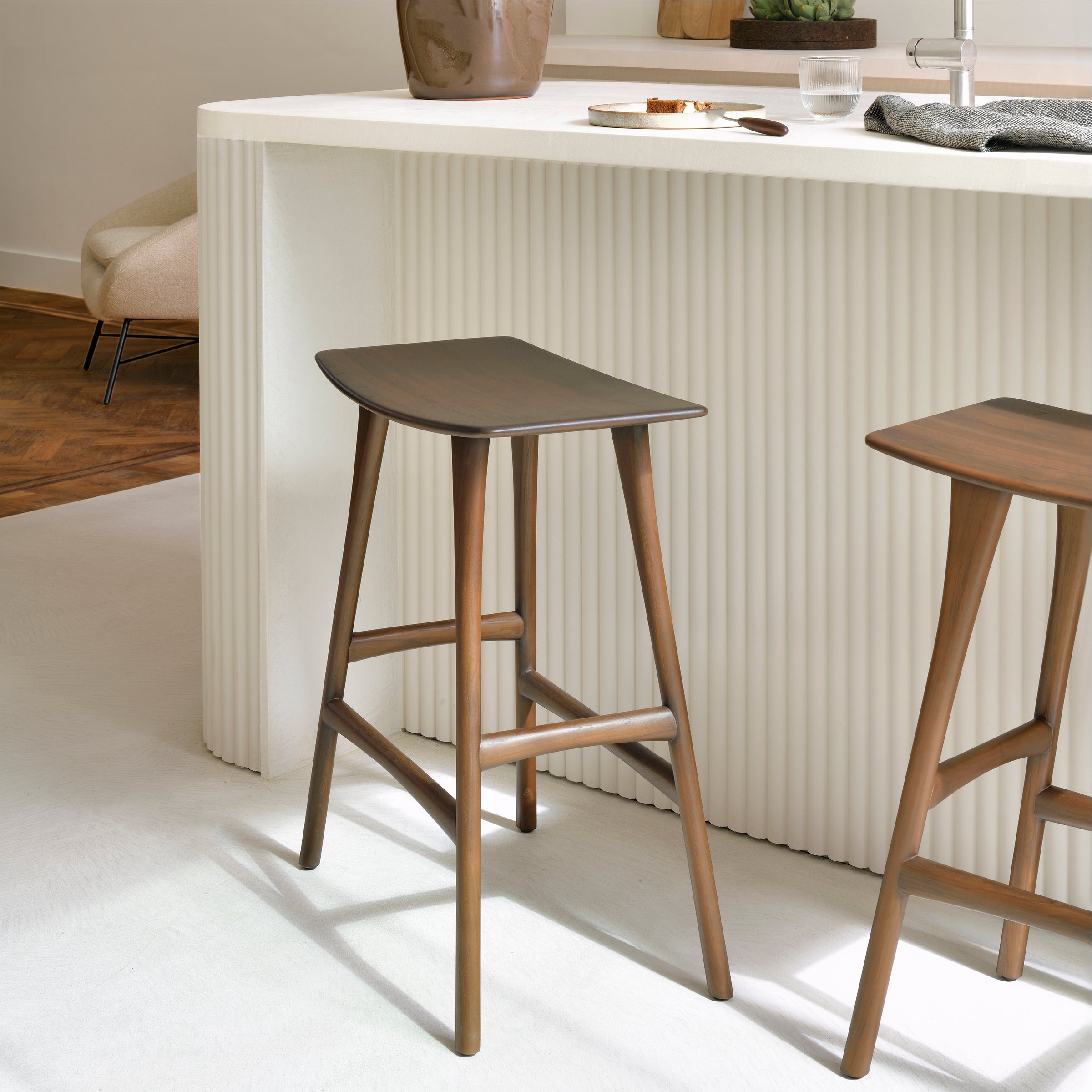 Osso Counter Stool in varnished teak with black leather upholstery placed next to a white kitchen island.