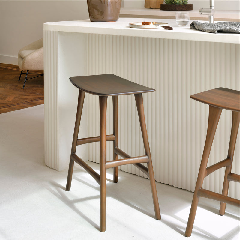 Osso Counter Stool in varnished teak with black leather upholstery placed next to a white kitchen island.