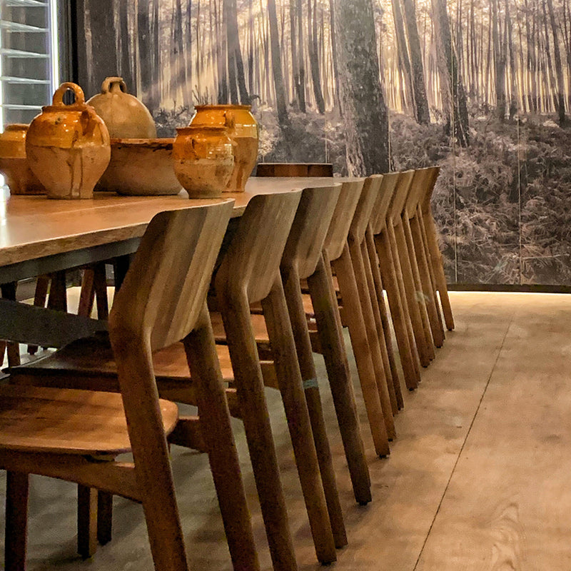 The dining area features a long solid wood table with matching FIN Chair Upholstered by Artisan. Large earthenware pots rest on the table, and a wall-sized forest scene photograph creates a natural, serene ambiance with its environmentally-friendly materials.