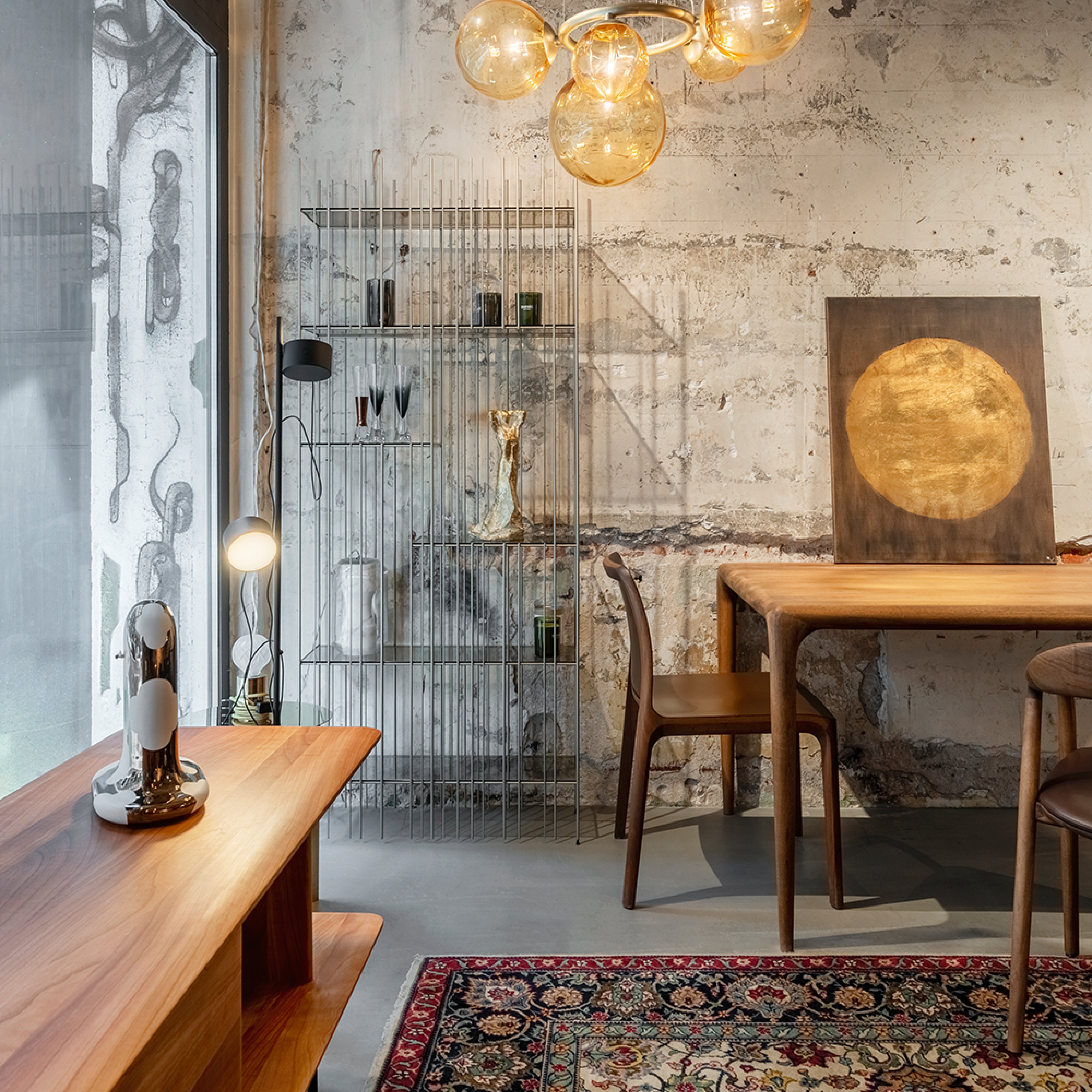 A modern room highlights an Artisan FIN Chair, a solid wood table and chairs set, a metal shelf with decor, and a warm-toned rug. A unique light fixture with round bulbs hangs above textured rustic walls. A window and wooden bench complete the left side of the room.
