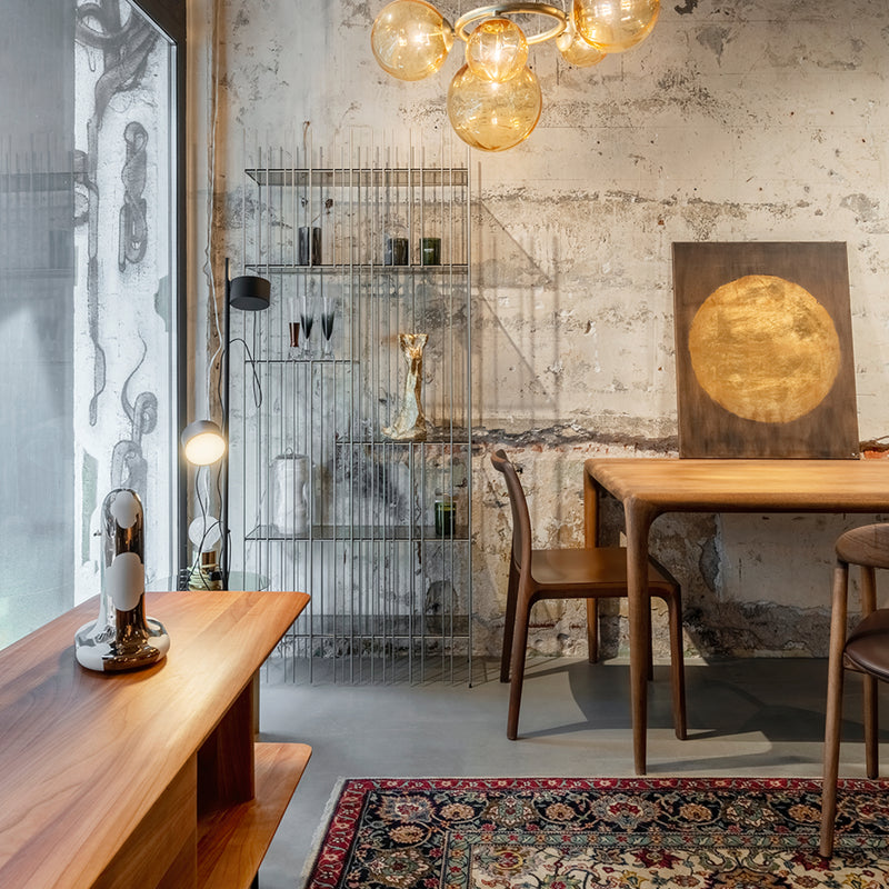 A stylish room features the FIN Chair Upholstered by Artisan, a solid wood table set, a metal shelf with decor, and a modern lamp. An abstract painting leans against the textured wall, while a colorful rug brightens part of the concrete floor beneath pendant lights.