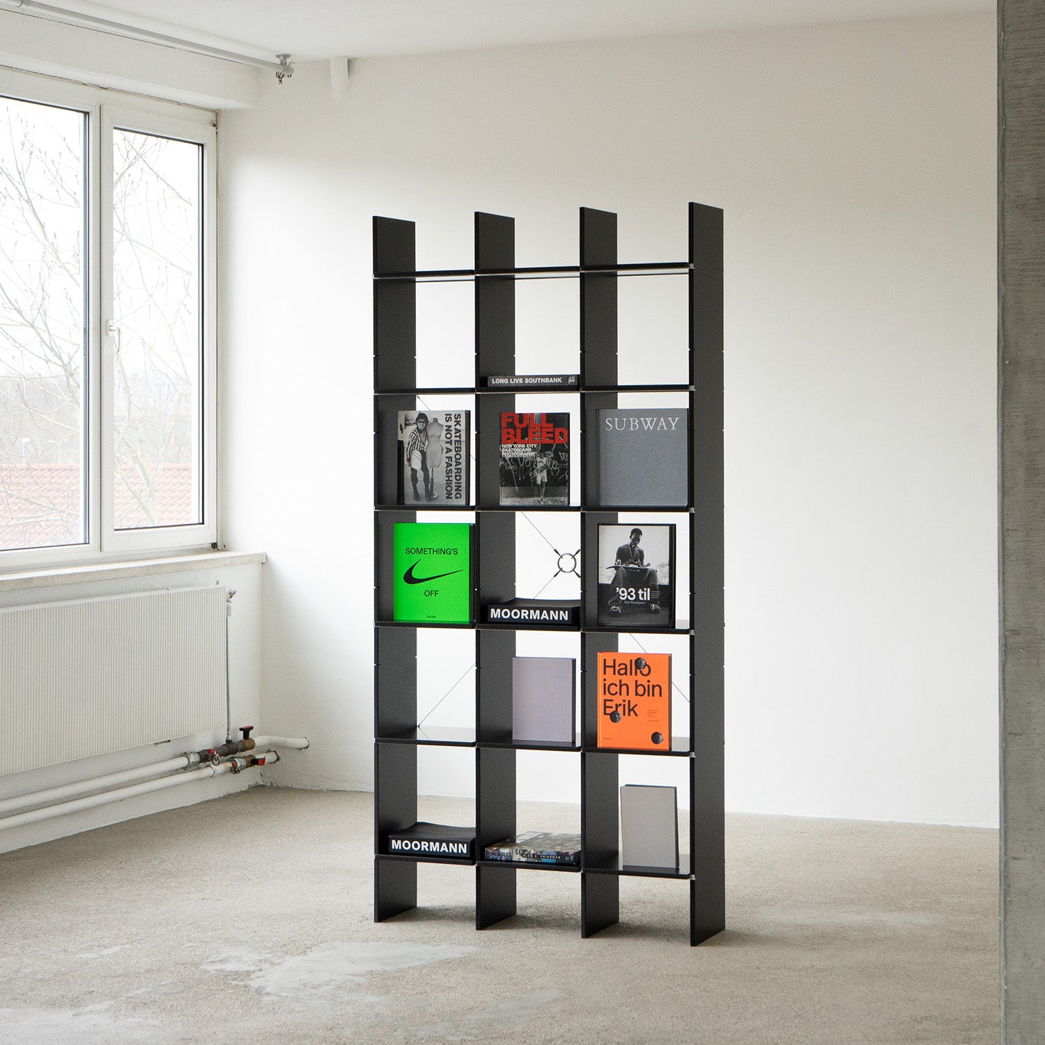 The modern black FNP shelf by Moormann features tall vertical dividers, ideal for holding books and magazines. Its design complements a minimalist room with a large window and white walls, enhancing a bright and airy atmosphere.