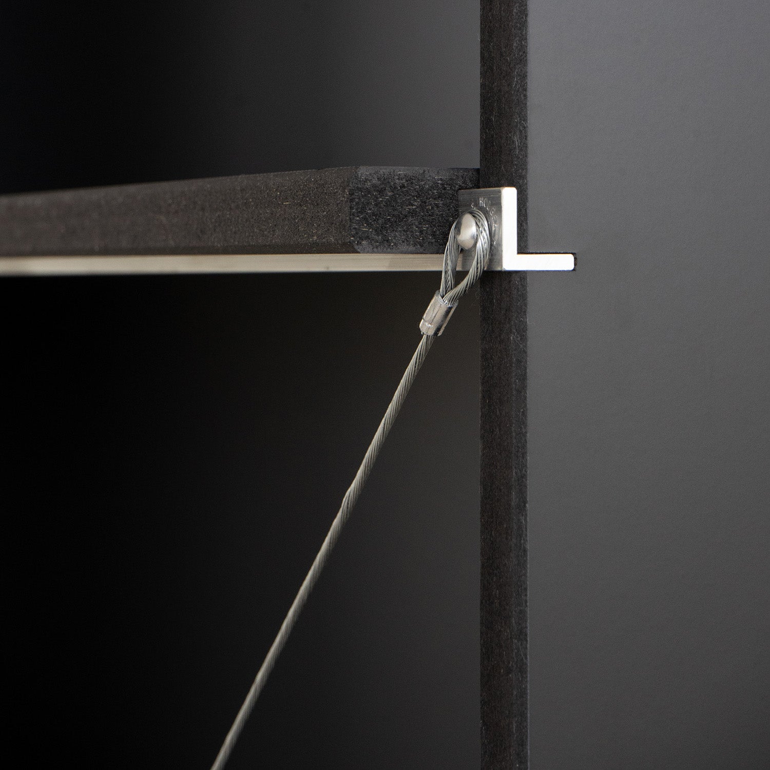 A close-up shows a tension cable affixed to a metal bracket on the edge of Moormanns FNP shelf against a dark backdrop, showcasing its minimalist and adaptable design with sleek, industrial materials.