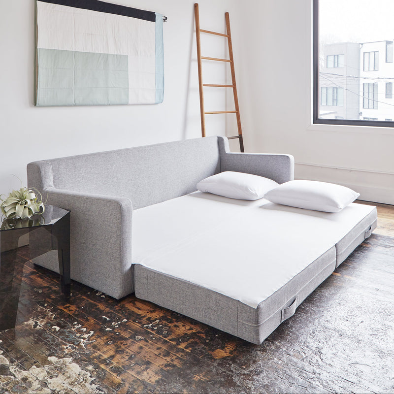 The Gus Modern Flipside Sofa Bed unfolds with white bedding and two pillows on a worn wooden floor in a bright room, featuring eco-friendly cushions, wall art, a ladder, window, and a small table with a plant.
