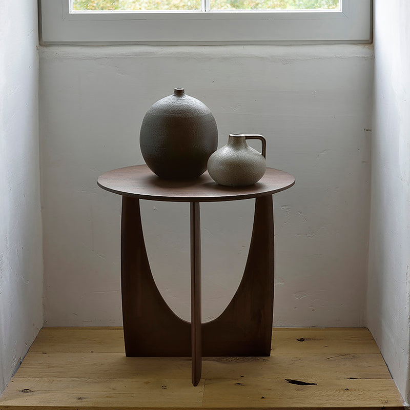 Ethnicraft’s Geometric Side Table, round with a sculptural base, holds two ceramic vases—one large and round, one small with a handle—displayed by a white wall and window above on a light wood floor.