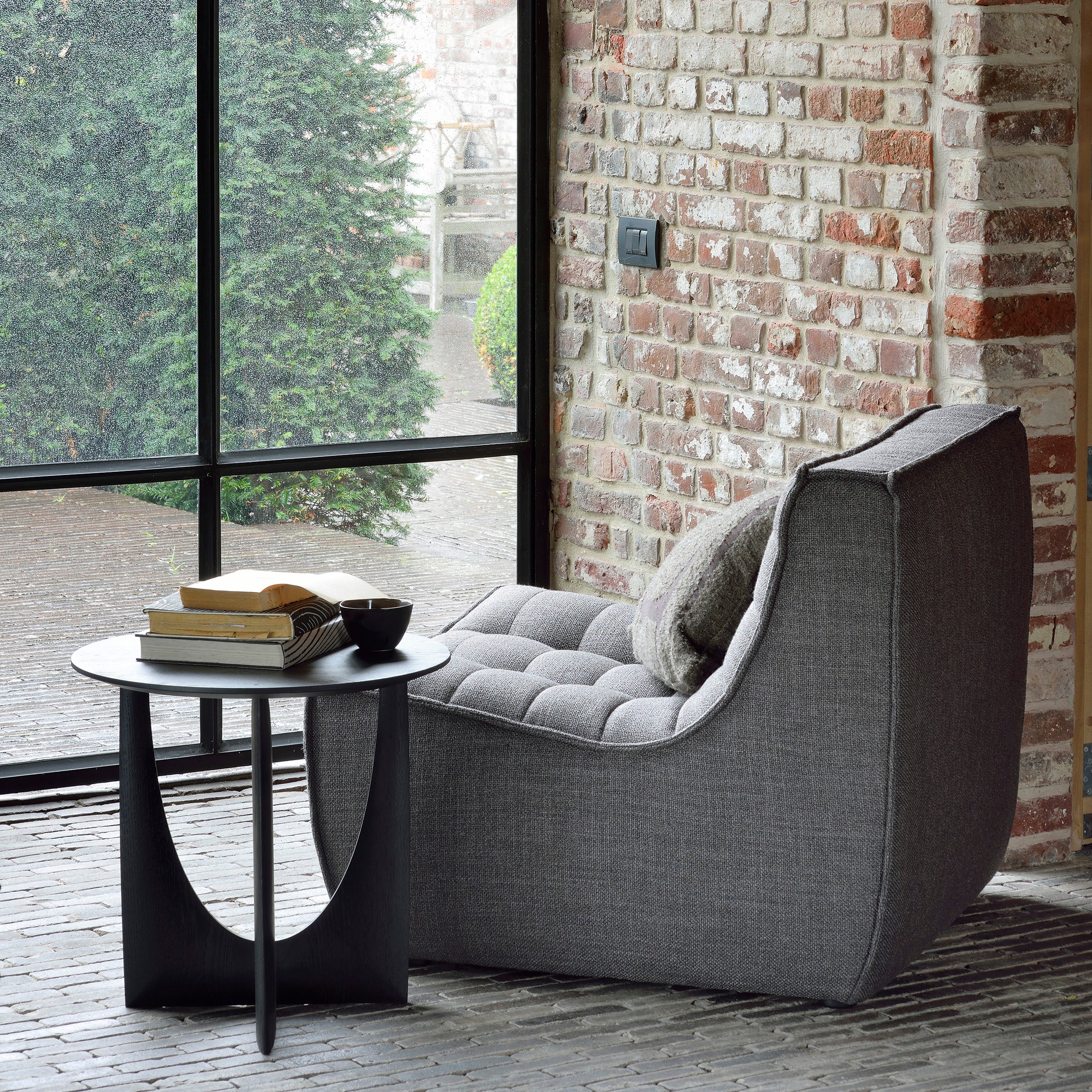 An Ethnicraft Geometric Side Table holds books and a cup beside a modern lounge chair with cushion, set by a large window and exposed brick wall overlooking greenery.