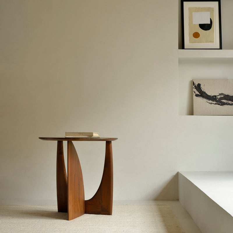 A minimalist room features the Ethnicraft Geometric Side Table with a book, set by a light wall. Built-in shelves above display abstract art and a canvas, enhancing the calm, modern vibe.