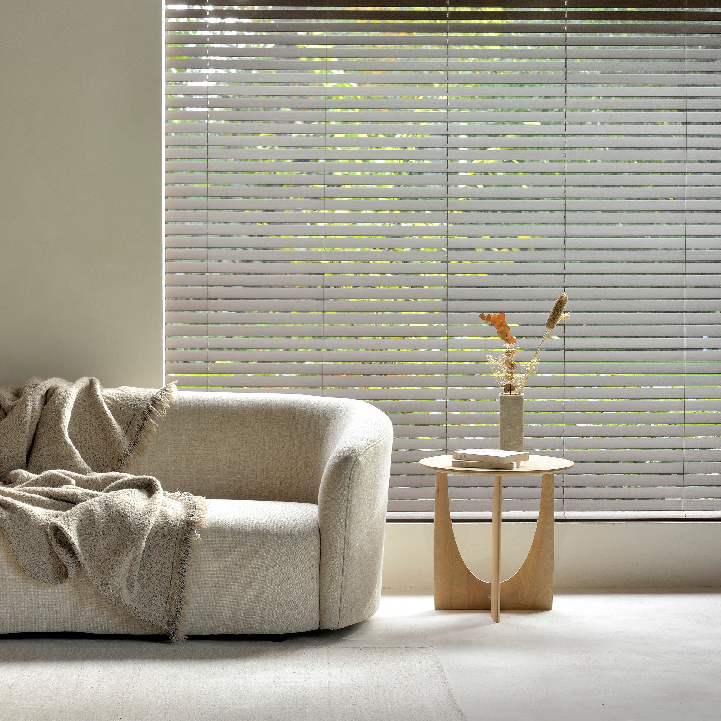 A modern, minimalist living room features a curved beige sofa with a textured throw, an Ethnicraft Geometric Side Table holding a vase of dried flowers, and large window blinds filtering in soft daylight.