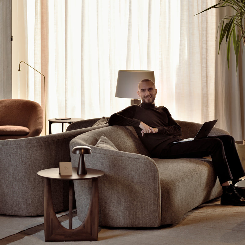 A man sits on a modern gray sofa with a laptop, smiling at the camera. The room is softly lit, featuring sheer curtains and an Ethnicraft Geometric Side Table, complemented by a lamp and an indoor plant.