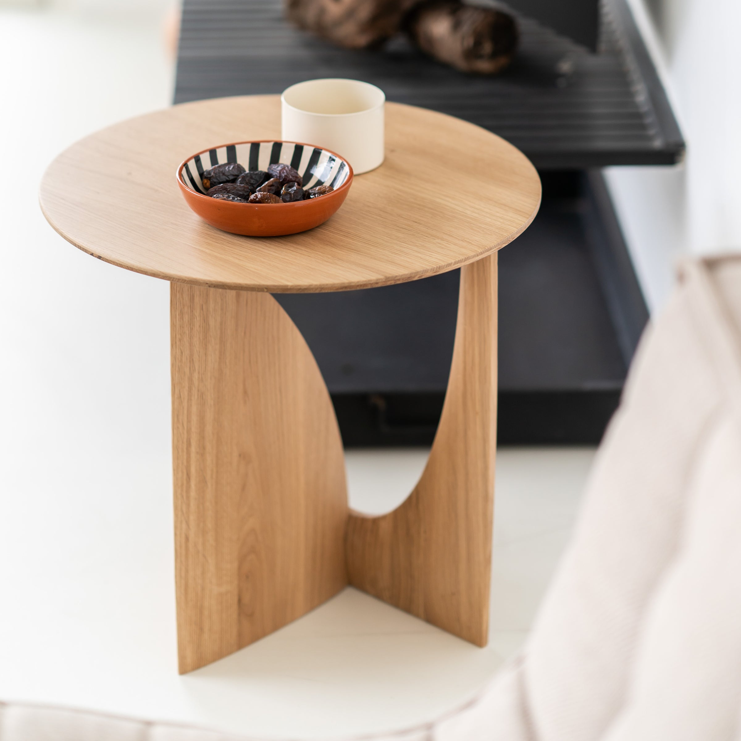 The Ethnicraft Geometric Side Table, a small and modern round piece, holds a striped bowl with dried fruit and a white cup. Placed beside a beige sofa, it sits near a black tray and other decorative accents in the background.