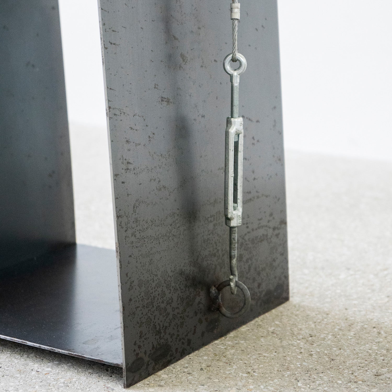 A close-up of a metal structure with a wire tensioning mechanism, featuring a turnbuckle and eye bolts, evokes the Gespanntes Regal by Moormann. The rust-textured surface highlights the steel cables durability, placed on a speckled concrete floor.