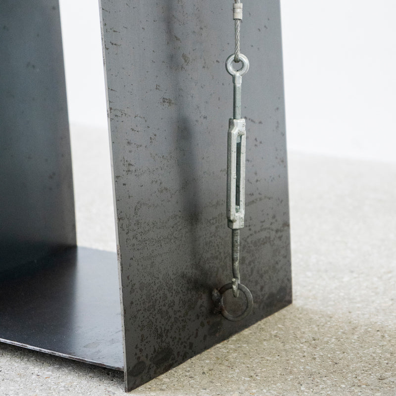 A close-up of a metal structure with a wire tensioning mechanism, featuring a turnbuckle and eye bolts, evokes the Gespanntes Regal by Moormann. The rust-textured surface highlights the steel cables durability, placed on a speckled concrete floor.